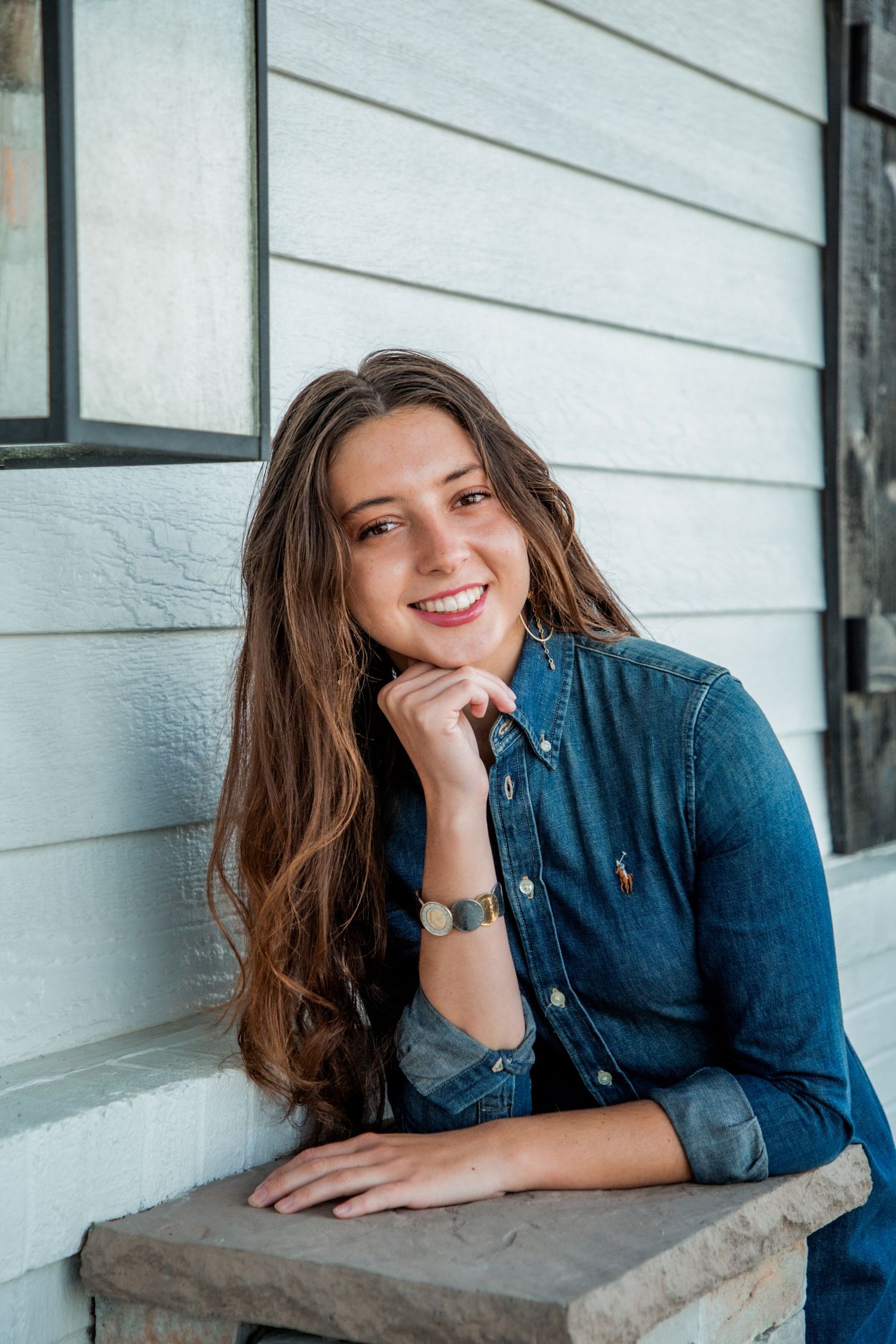 Young woman in denim shirt smiling warmly while resting chin on hand on white farmhouse porch.