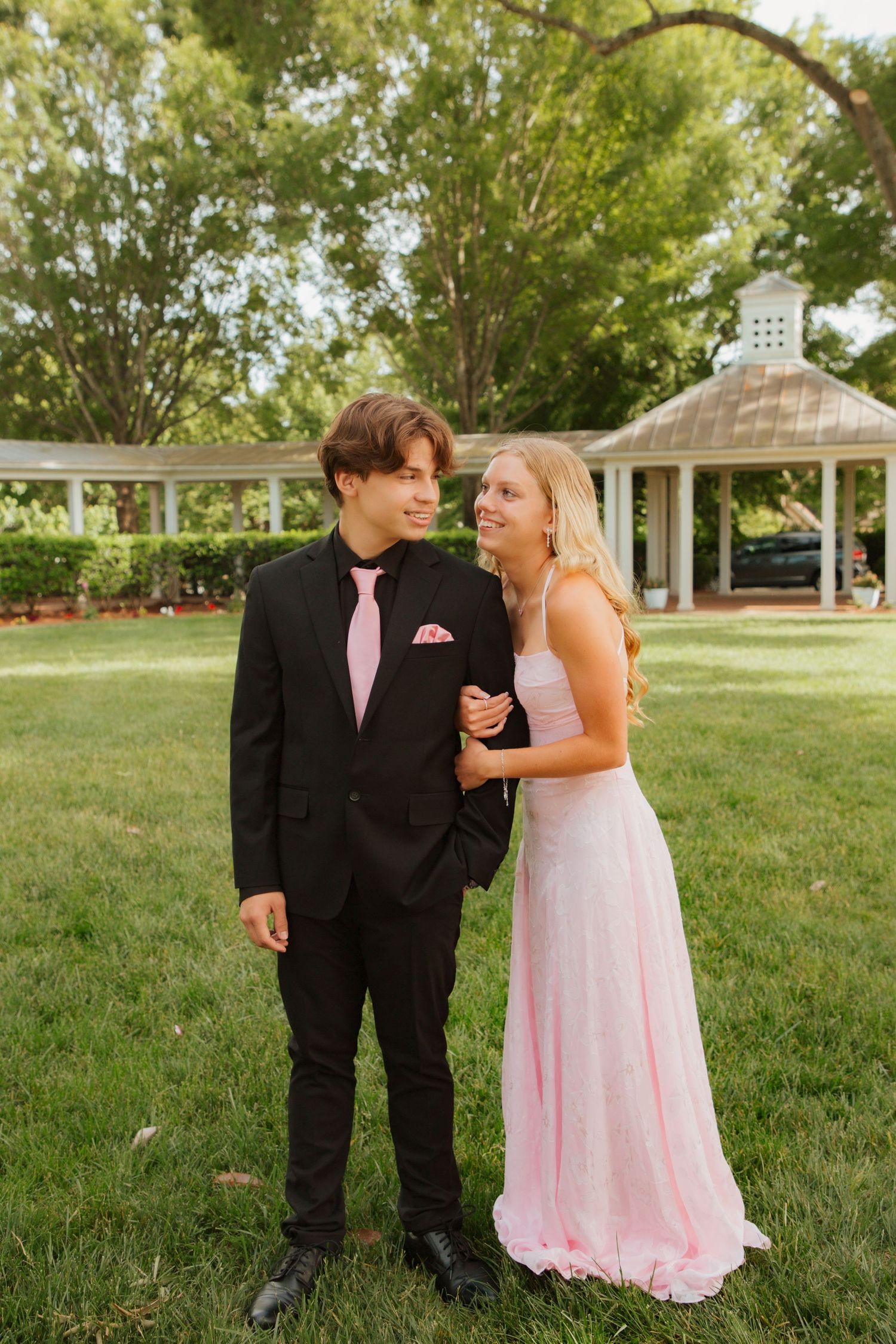 Smiling prom couple in black suit and pink gown stand together on lush green lawn.