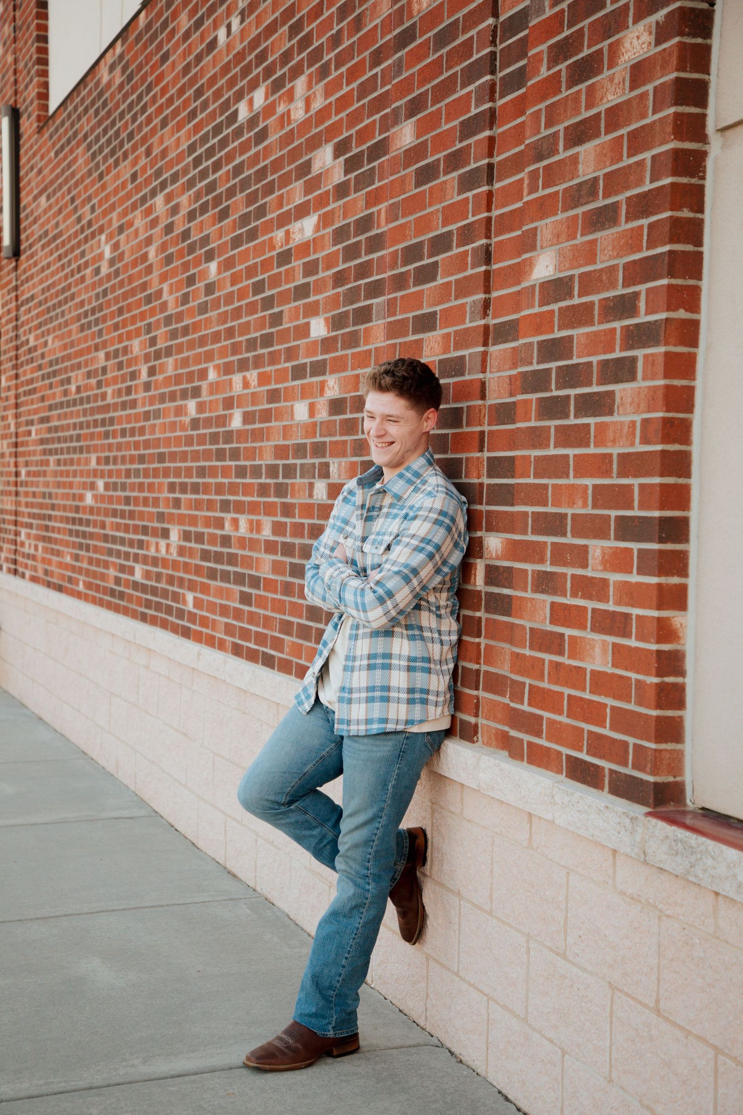 Teen boy in plaid shirt leaning against brick wall laughing during senior photos.