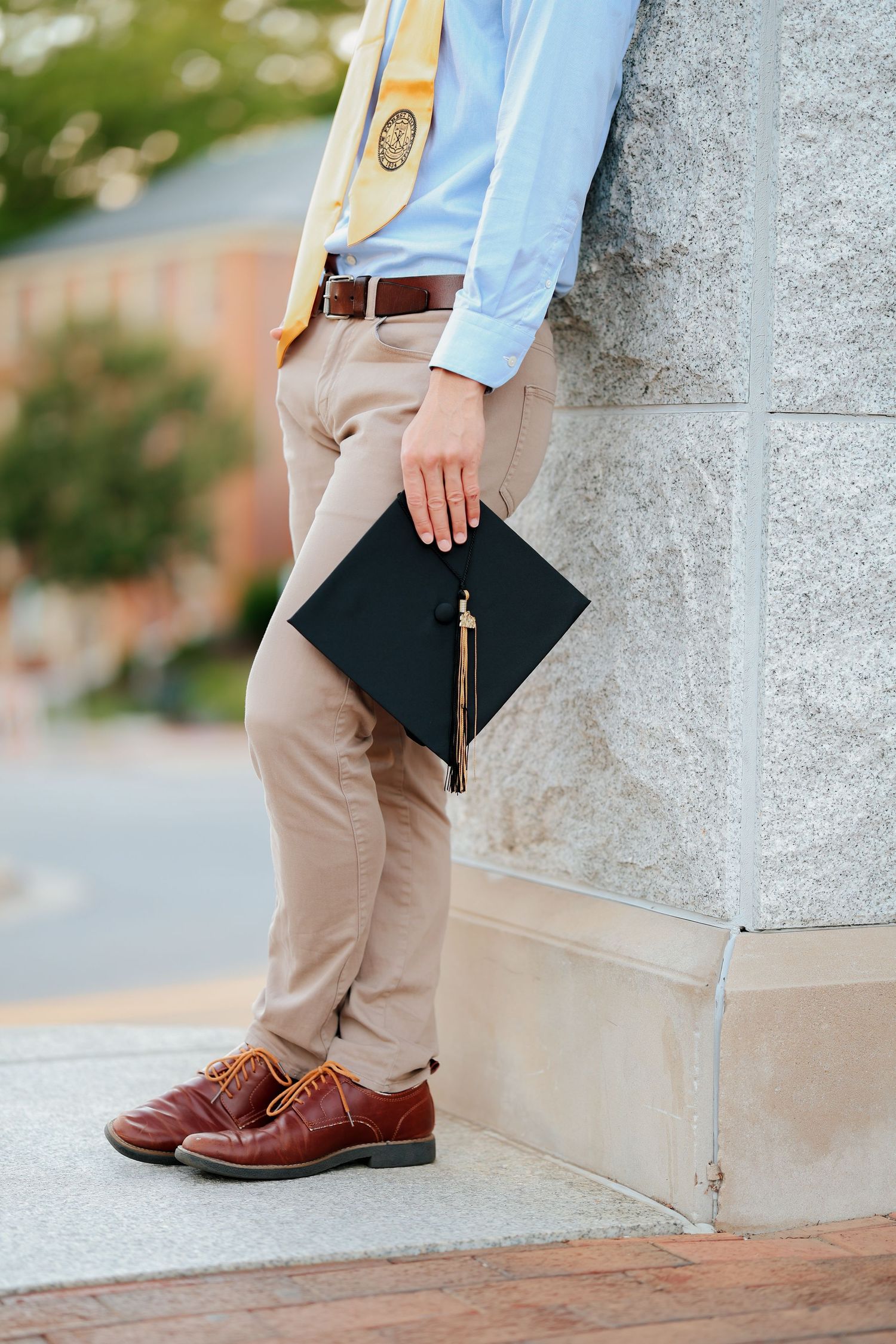 Graduate holding a black cap with gold tassel wearing khaki pants and brown dress shoes outdoors.