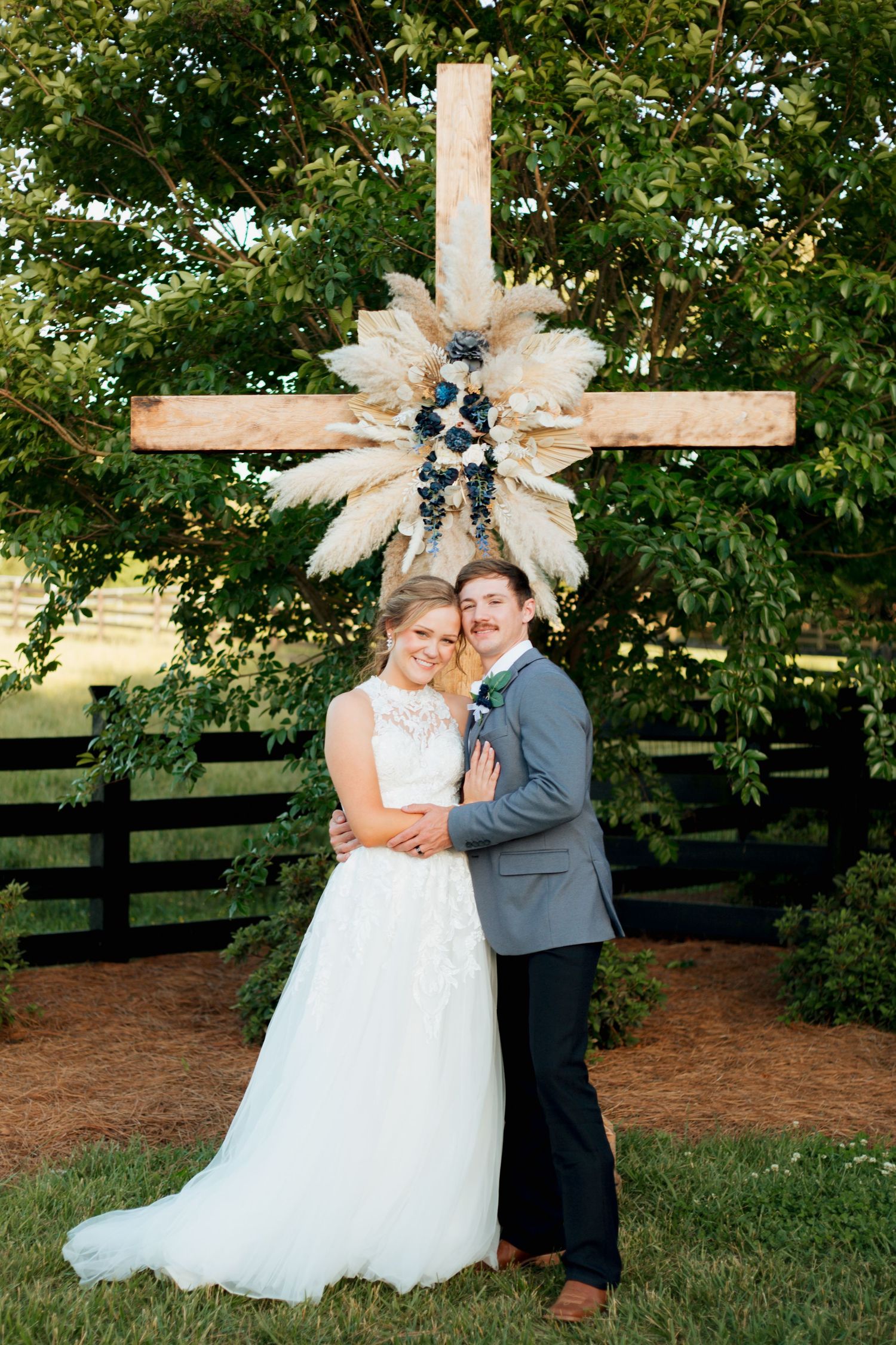 Bride and groom pose in front of wooden cross decorated with pampas grass.