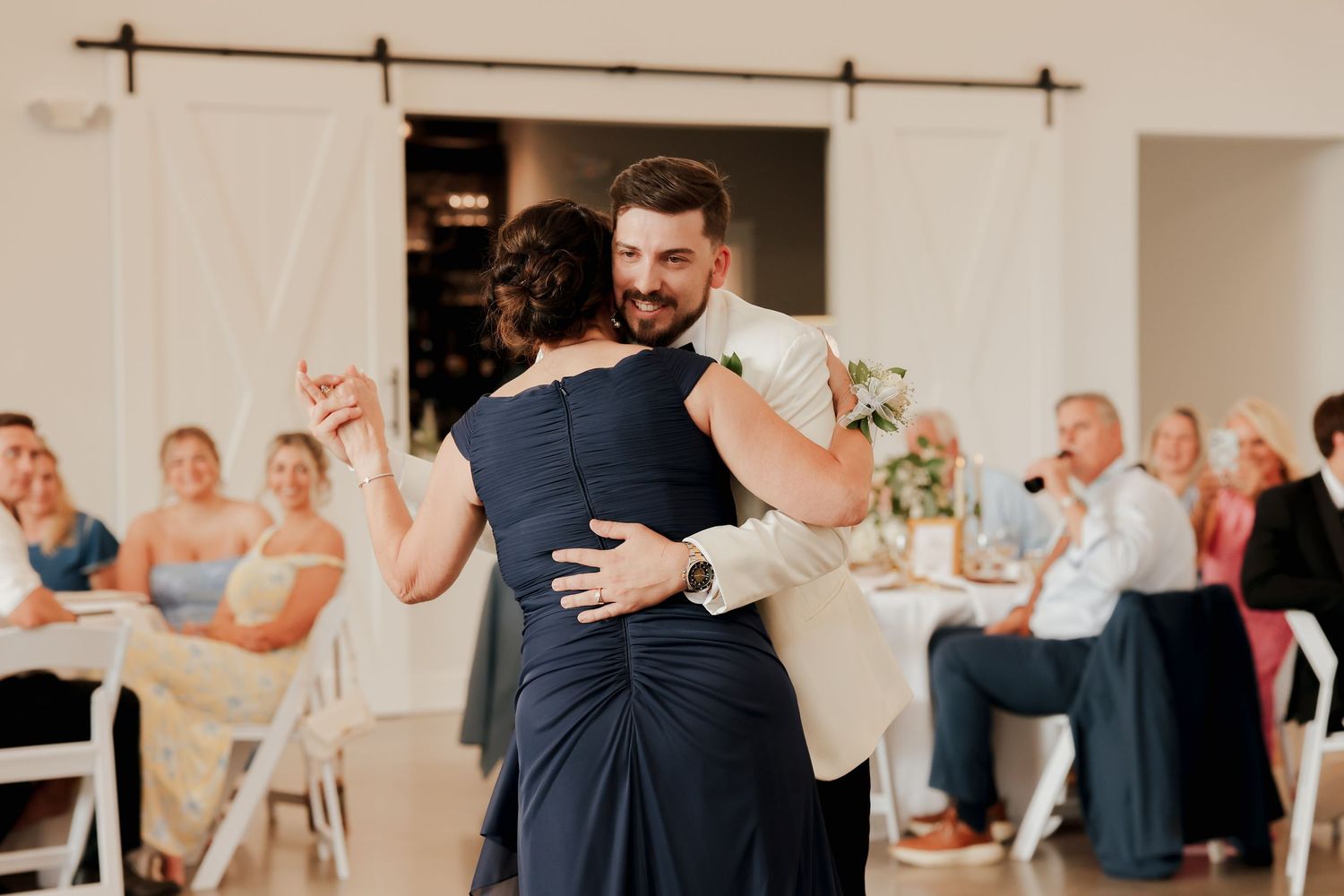 Groom in white tuxedo shares emotional mother-son dance at elegant wedding reception with barn doors.