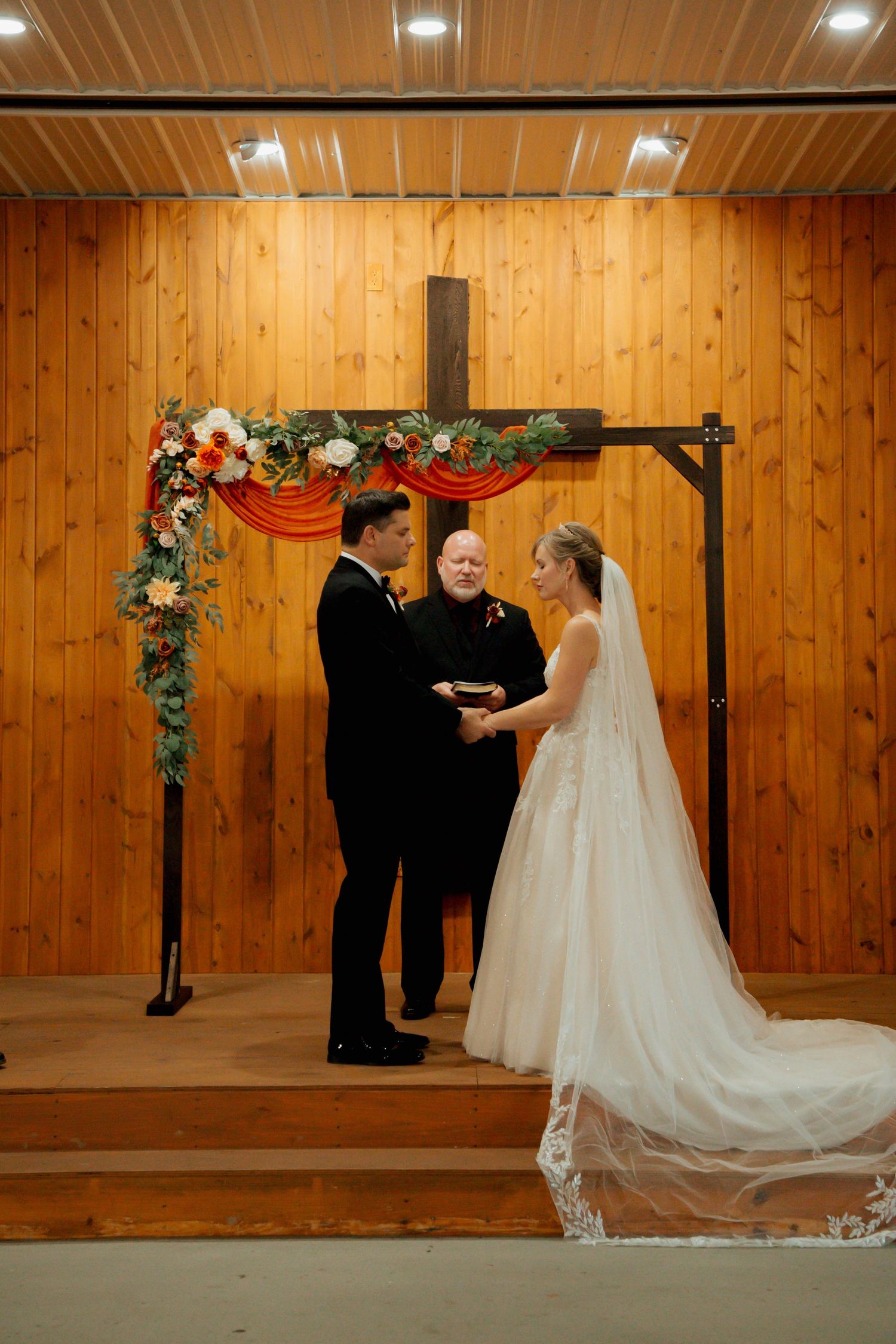 A bride and groom exchange vows beneath a floral arch with a wooden cross in a rustic chapel.