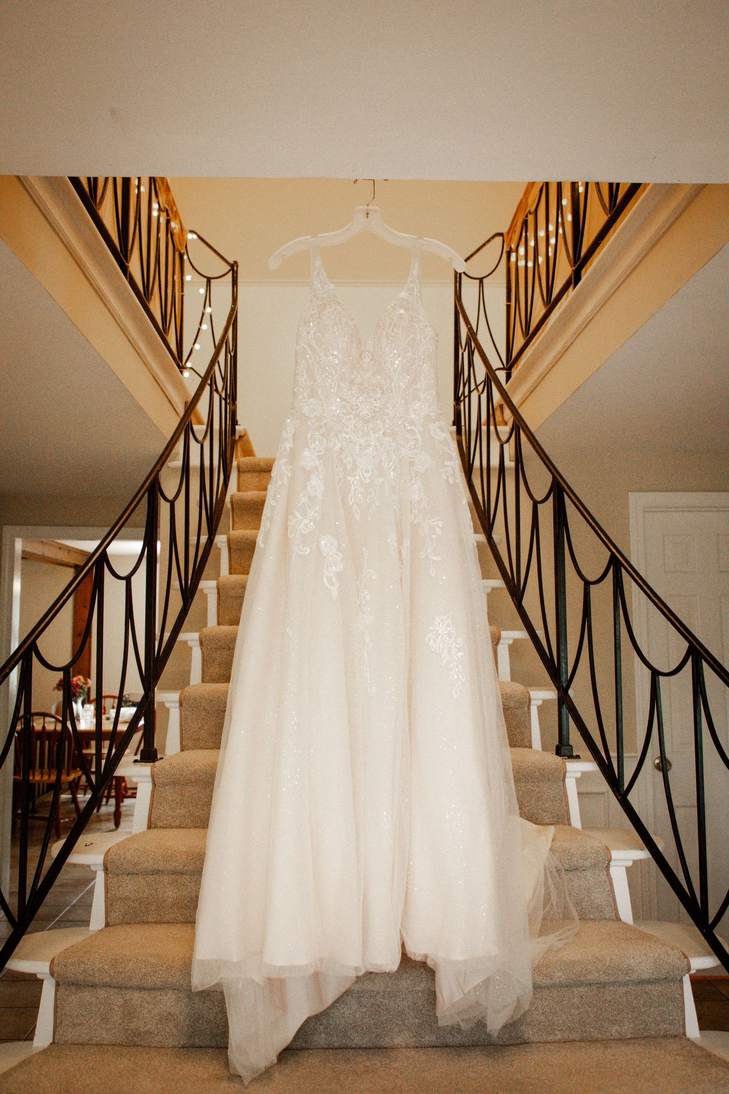 Elegant lace A-line wedding gown hanging on staircase railing in a grand home with wrought iron balusters.