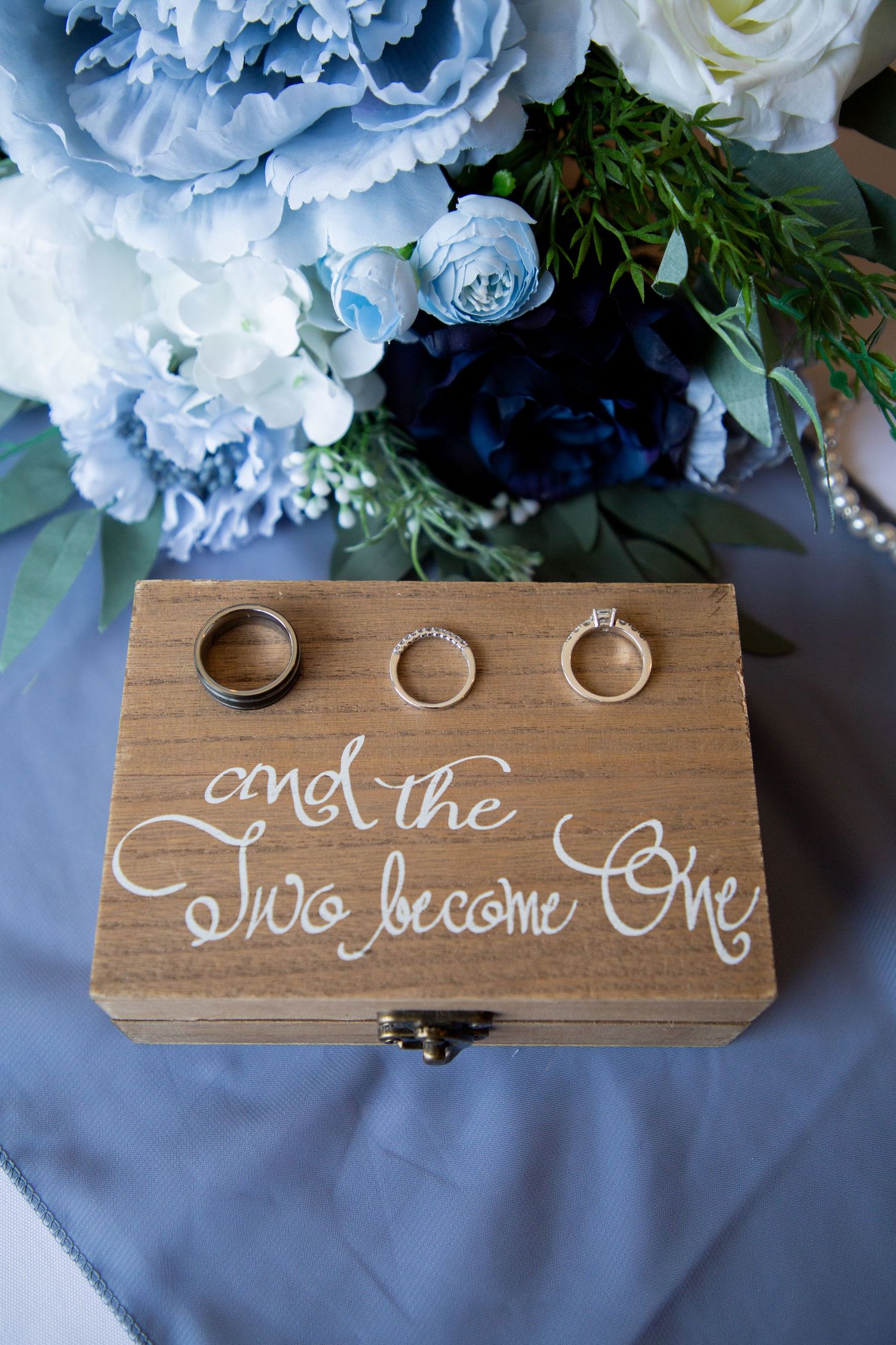 Three wedding rings on a wooden box inscribed with and the two become one beside a blue bouquet.