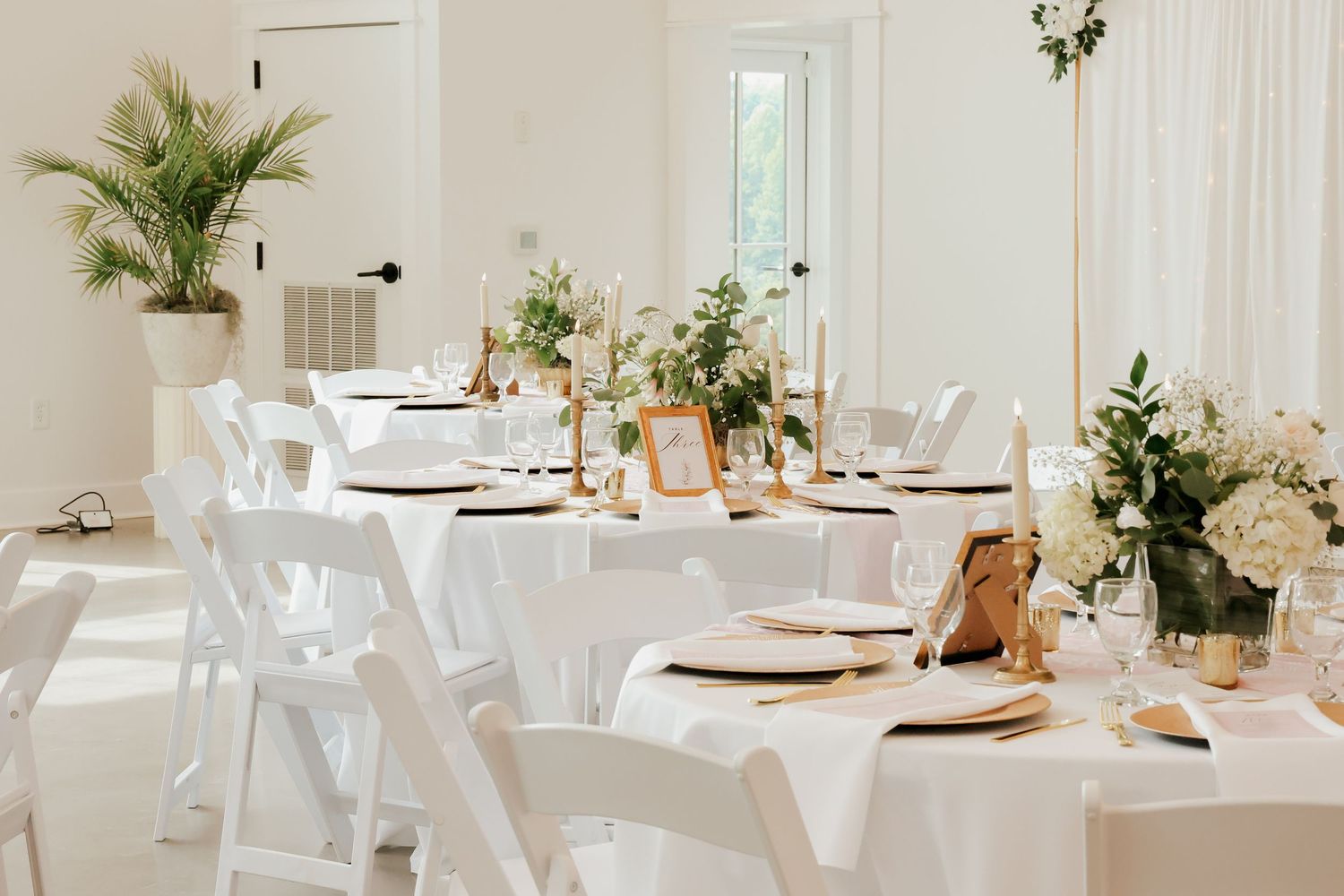 Elegant white wedding reception hall with long tables, gold candlesticks, and greenery centerpieces.