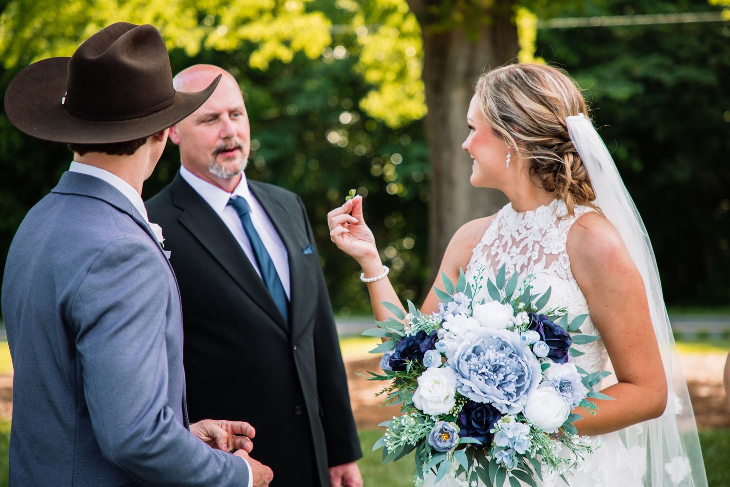 Bride holding navy and white bouquet exchanges vows with groom in gray suit and cowboy hat during outdoor wedding ceremony.