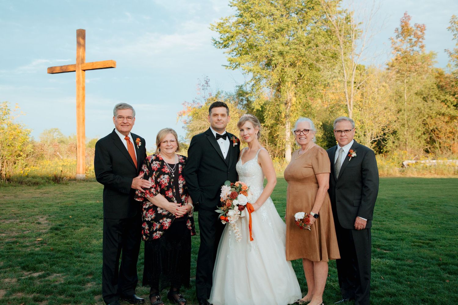 Bride and groom with four family members posing near a wooden cross at an outdoor wedding.