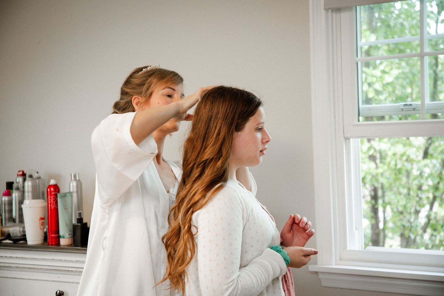 Bridesmaid styling bride's long red hair before wedding ceremony near window.