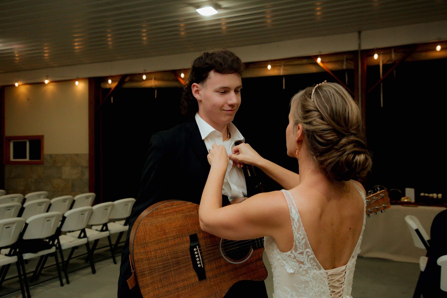 Bride in lace gown adjusting groom's tie near acoustic guitar in wedding venue.