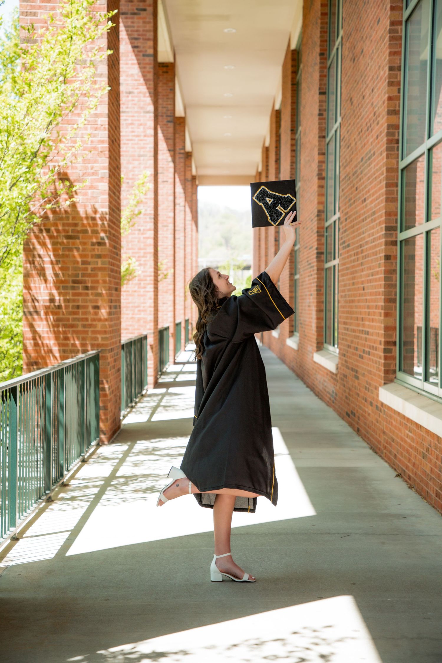 Graduate in black robe joyfully tossing cap in brick building corridor.