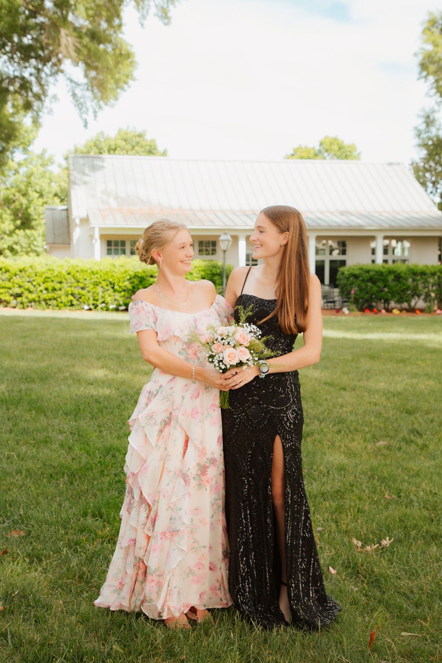 Two teenage girls in formal dresses, one floral pink and one black sequin, pose on green lawn.