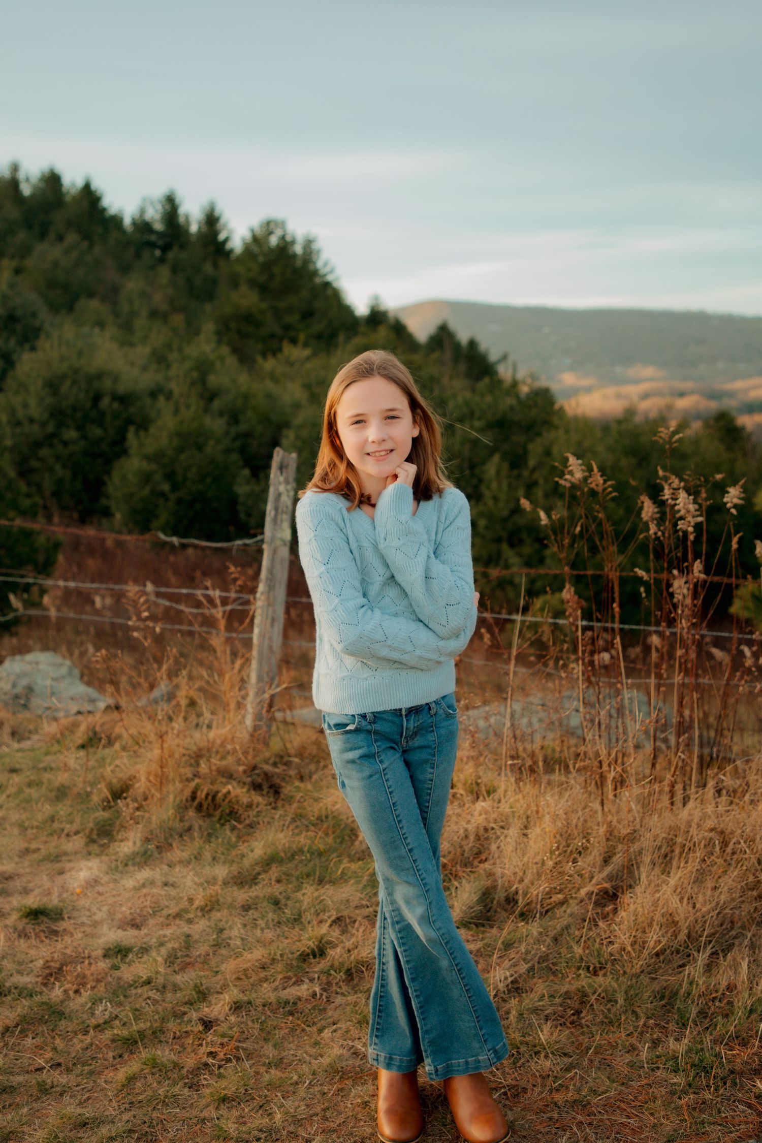 Young girl in blue sweater and flare jeans poses thoughtfully in a golden field with mountain backdrop.
