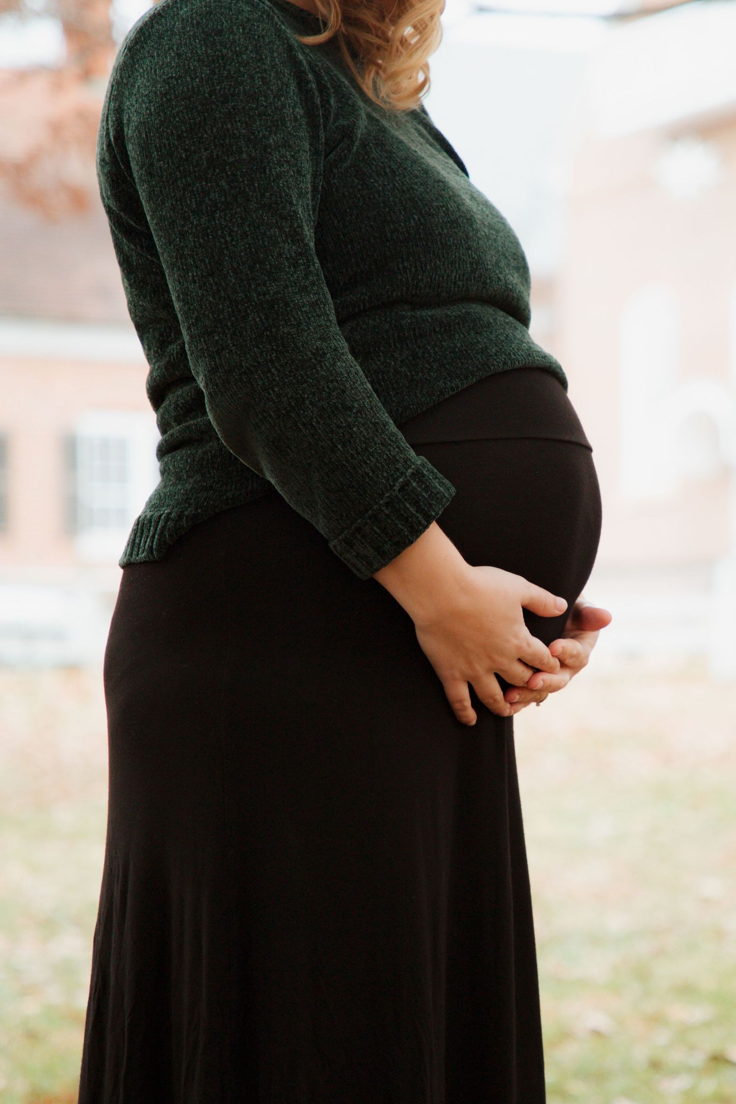Close-up side profile of pregnant woman in green sweater cradling baby bump outdoors.