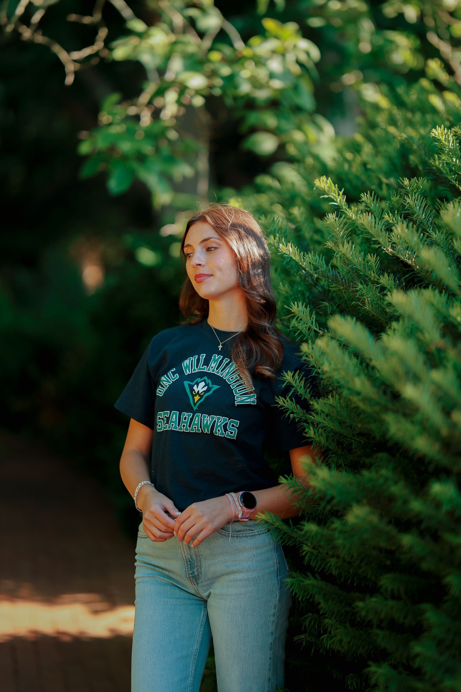 Young woman in UNCW Seahawks shirt standing beside evergreen trees in dappled sunlight.