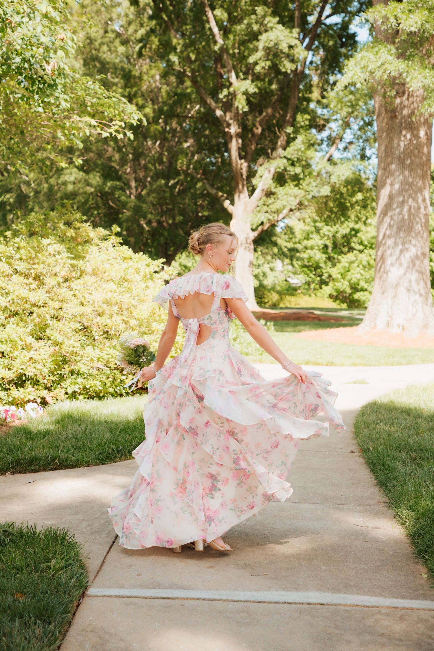 A young woman twirls in a floral pink dress on a garden path surrounded by lush green trees.