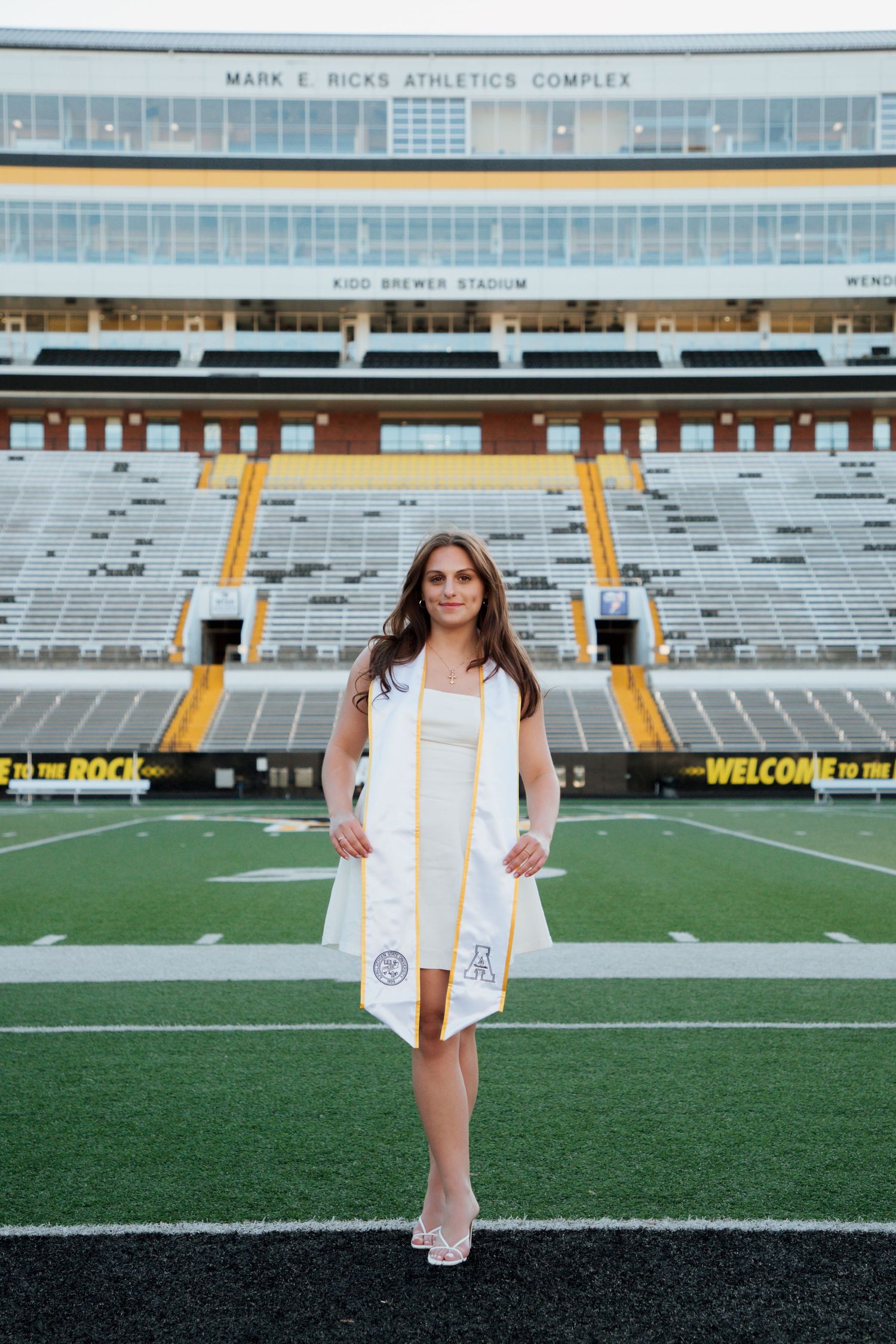 Young woman in white graduation gown and stole stands on football field at Kidd Brewer Stadium.