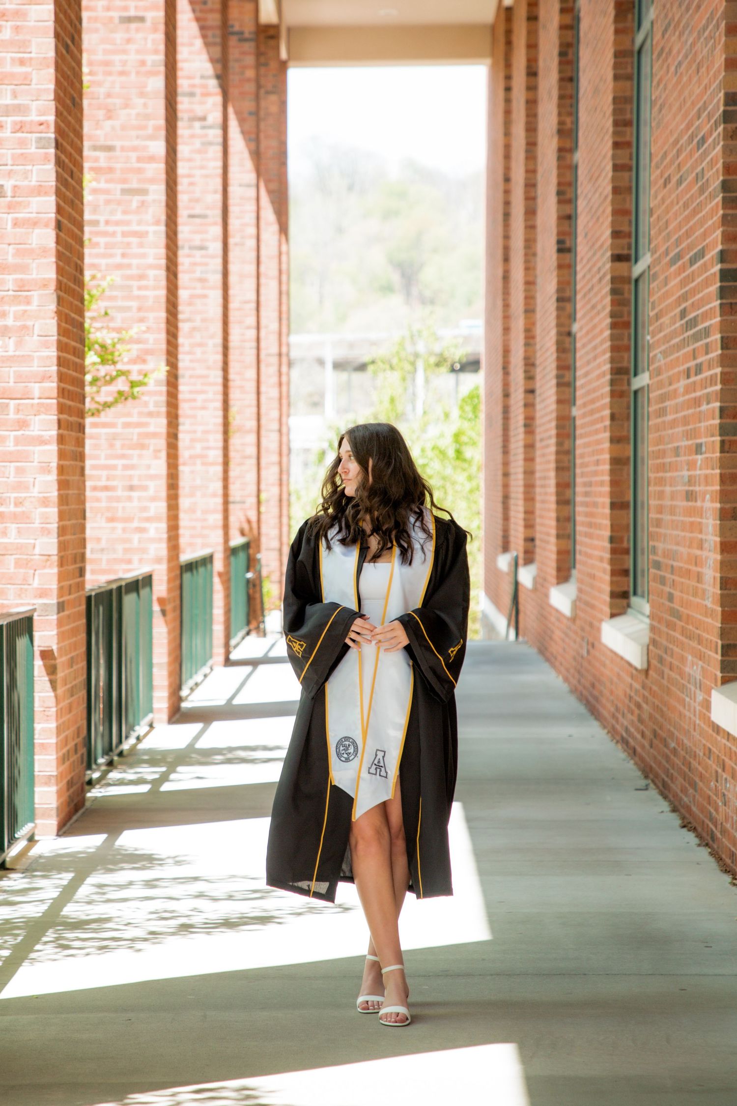 Graduate in black robe walking through sunlit brick corridor wearing white stole.