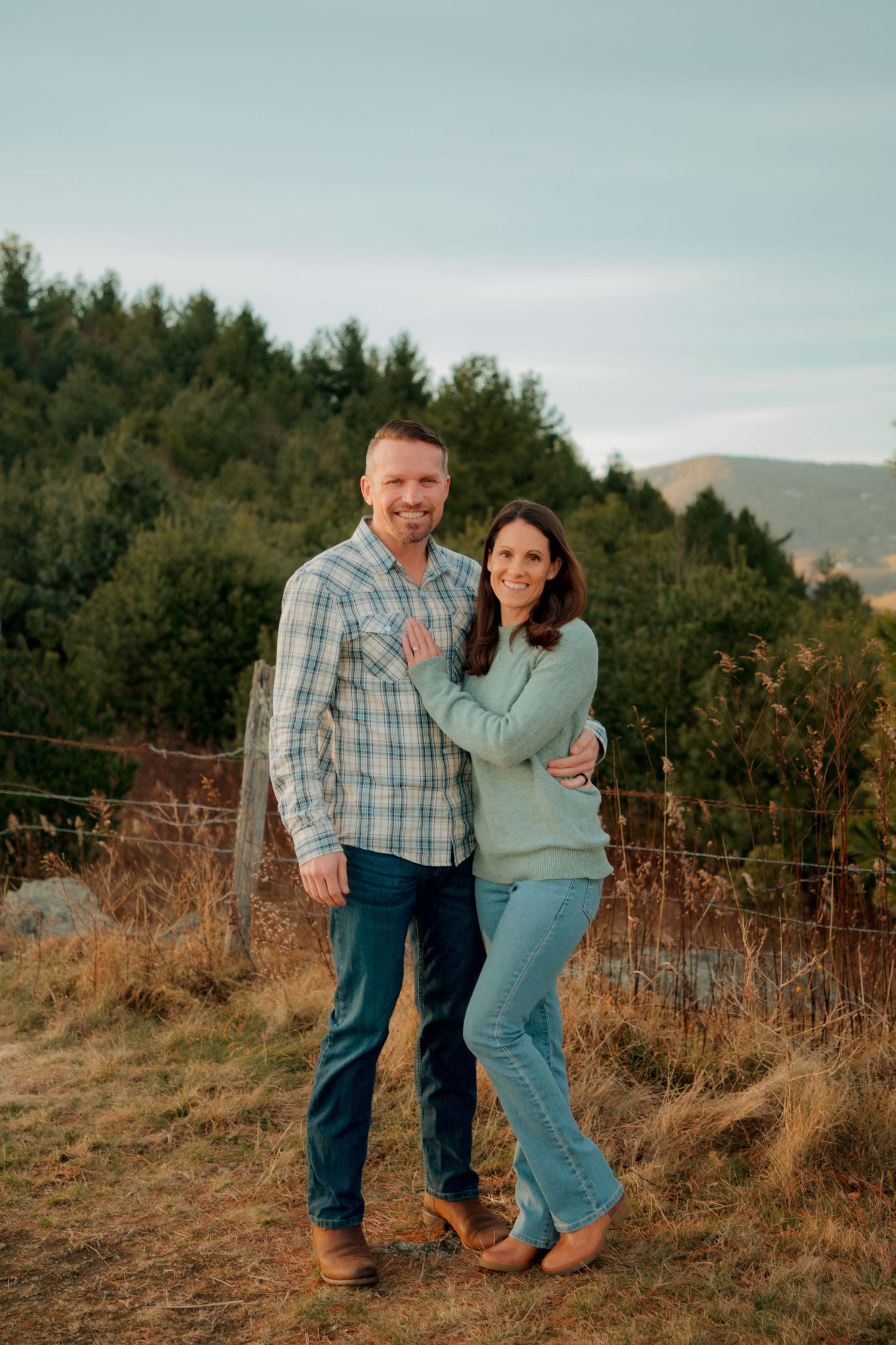 Smiling couple in coordinating blue outfits embraces outdoors with scenic mountain and forest background.