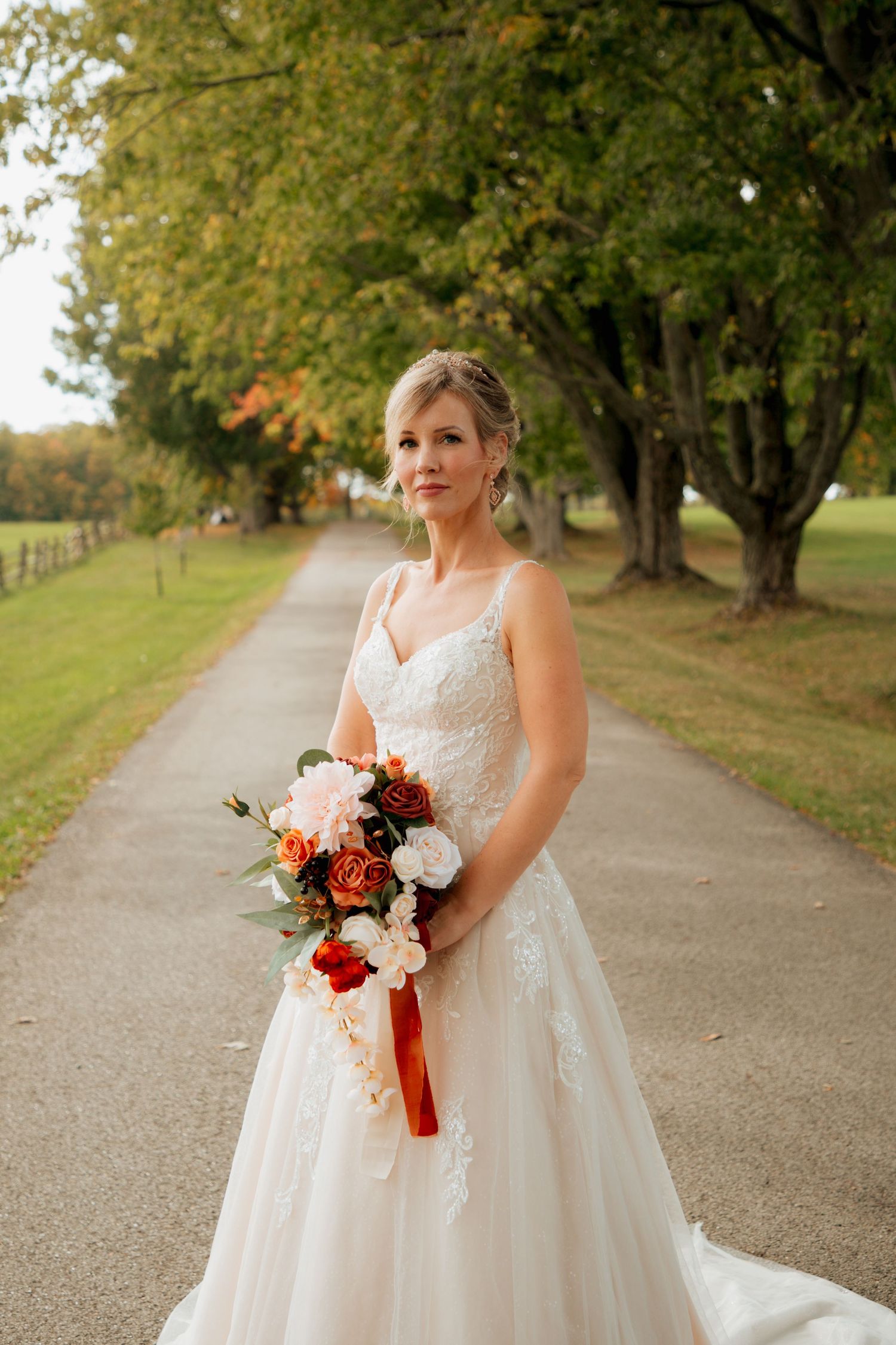 Beautiful bride in a lace A-line gown holds a stunning fall bouquet of red and white roses on a tree-lined path.