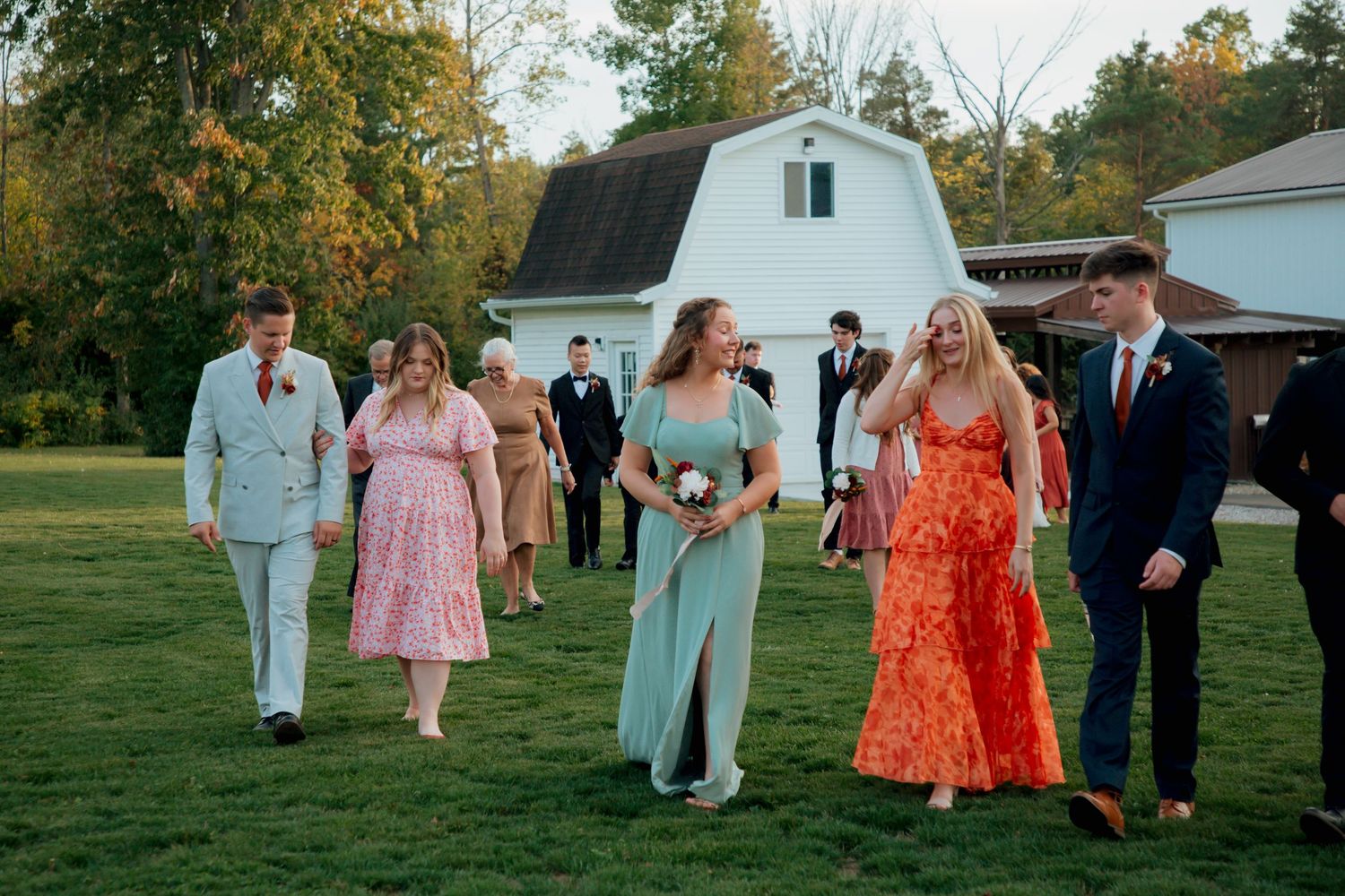 Wedding party guests walk across green lawn with white barn in background during outdoor autumn wedding celebration.