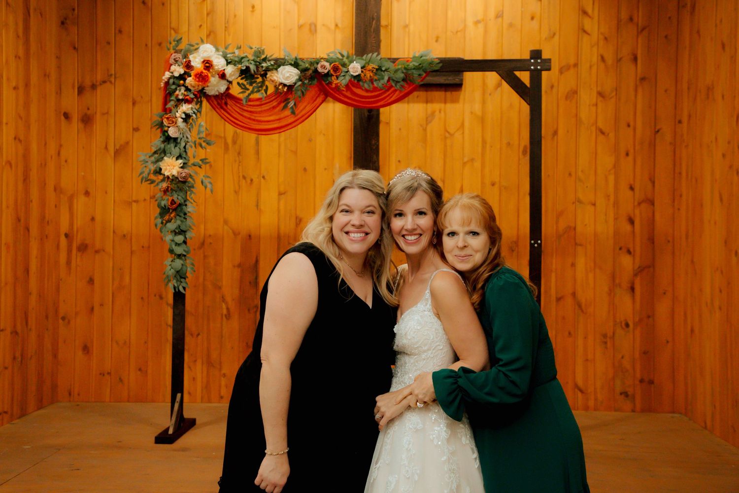 Bride posing with two women in front of decorated cross wedding arch in rustic venue.