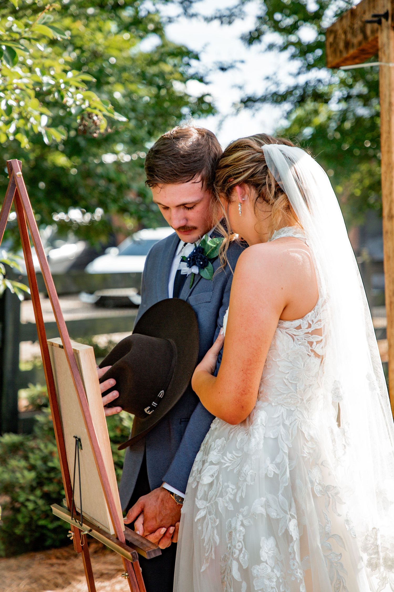 Bride and groom bow heads together in prayer at wooden easel during outdoor wedding ceremony.