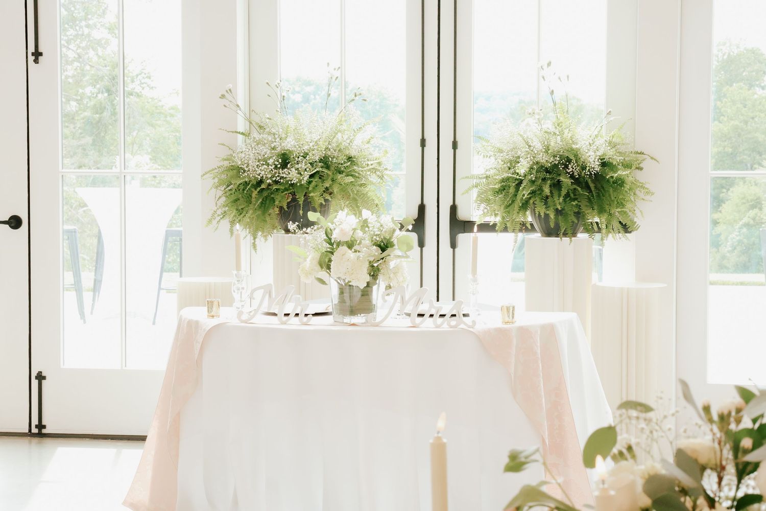 Elegant sweetheart table with white linen, greenery centerpieces, and candles by bright windows.
