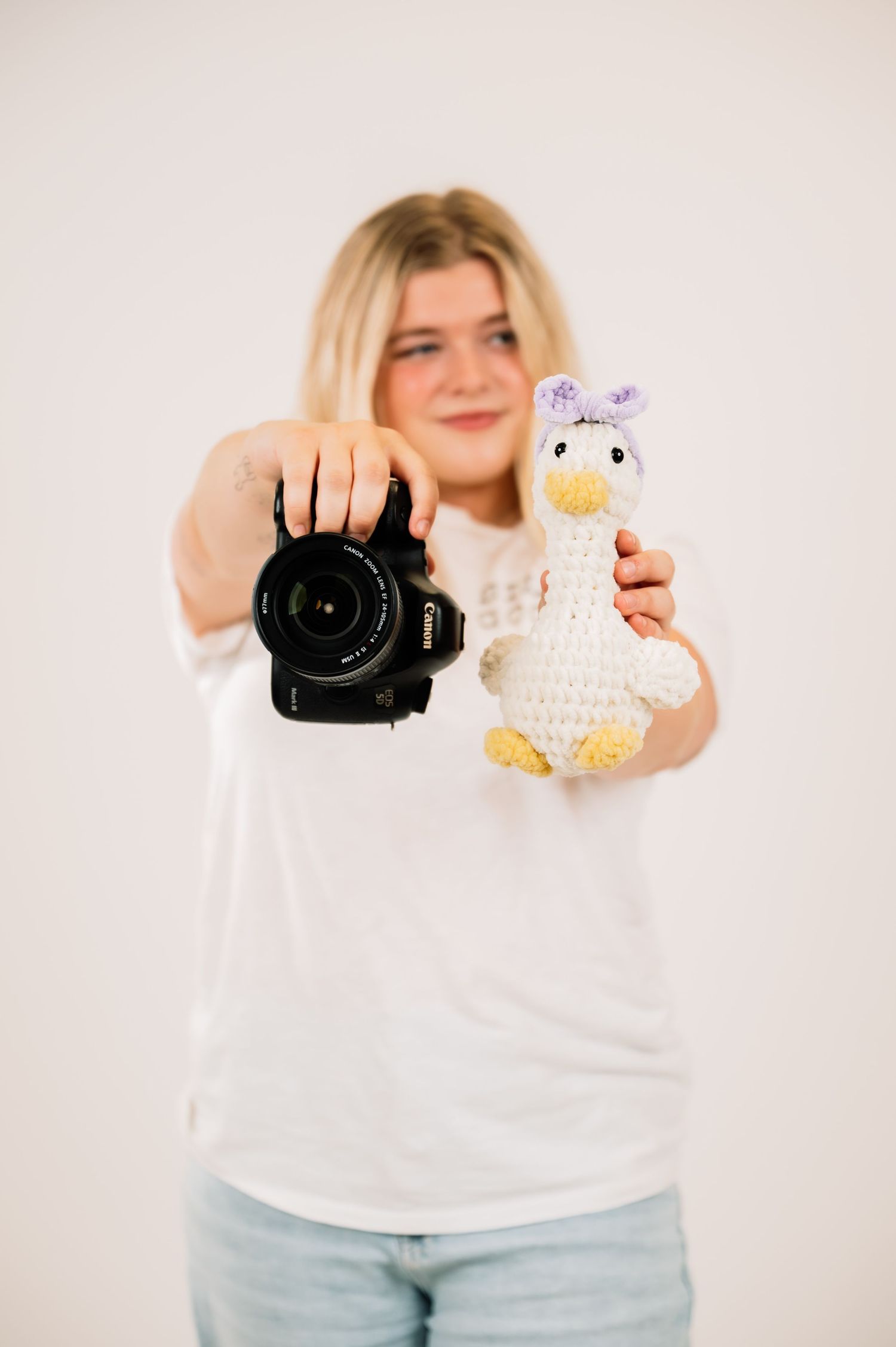 Blonde female photographer holding Canon DSLR camera and crocheted duck toy toward camera.