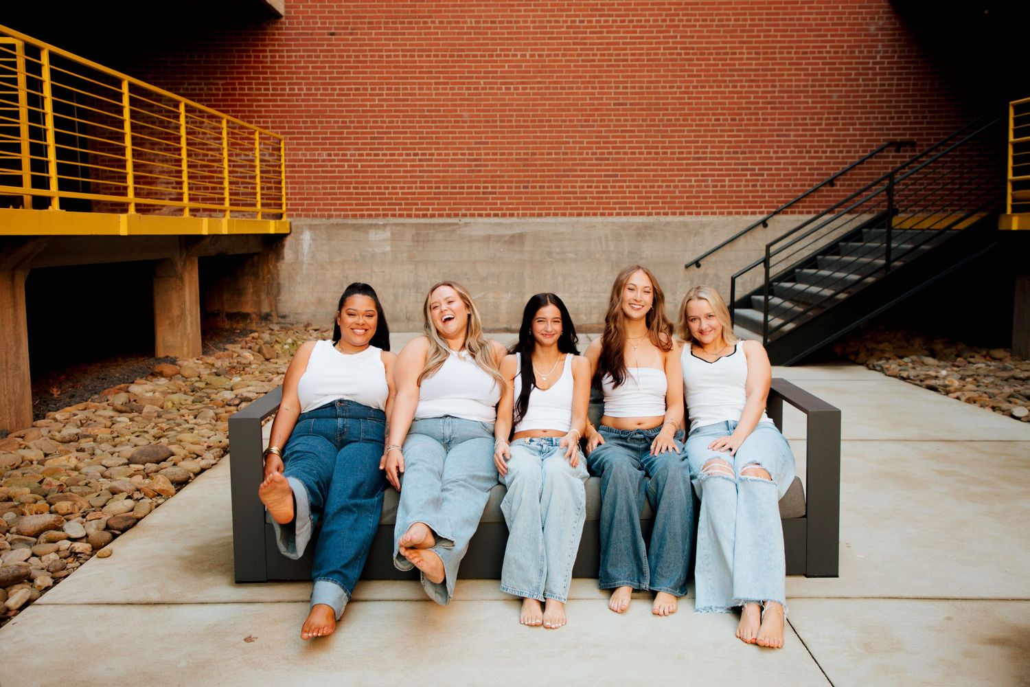 Five women in white tops and jeans sit together laughing on an outdoor bench against a brick wall.