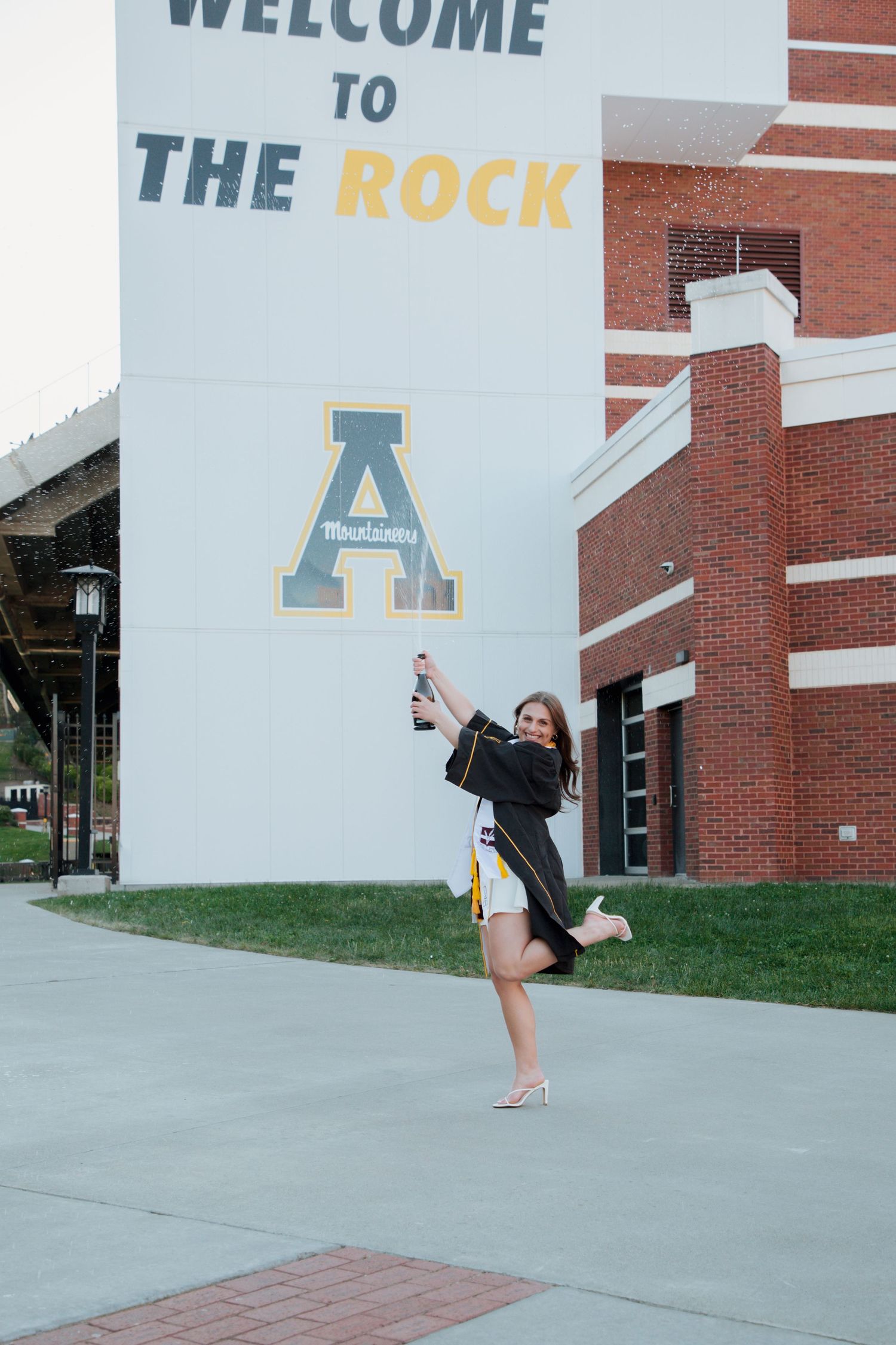 Joyful graduate popping champagne at Welcome to The Rock Appalachian State University stadium.