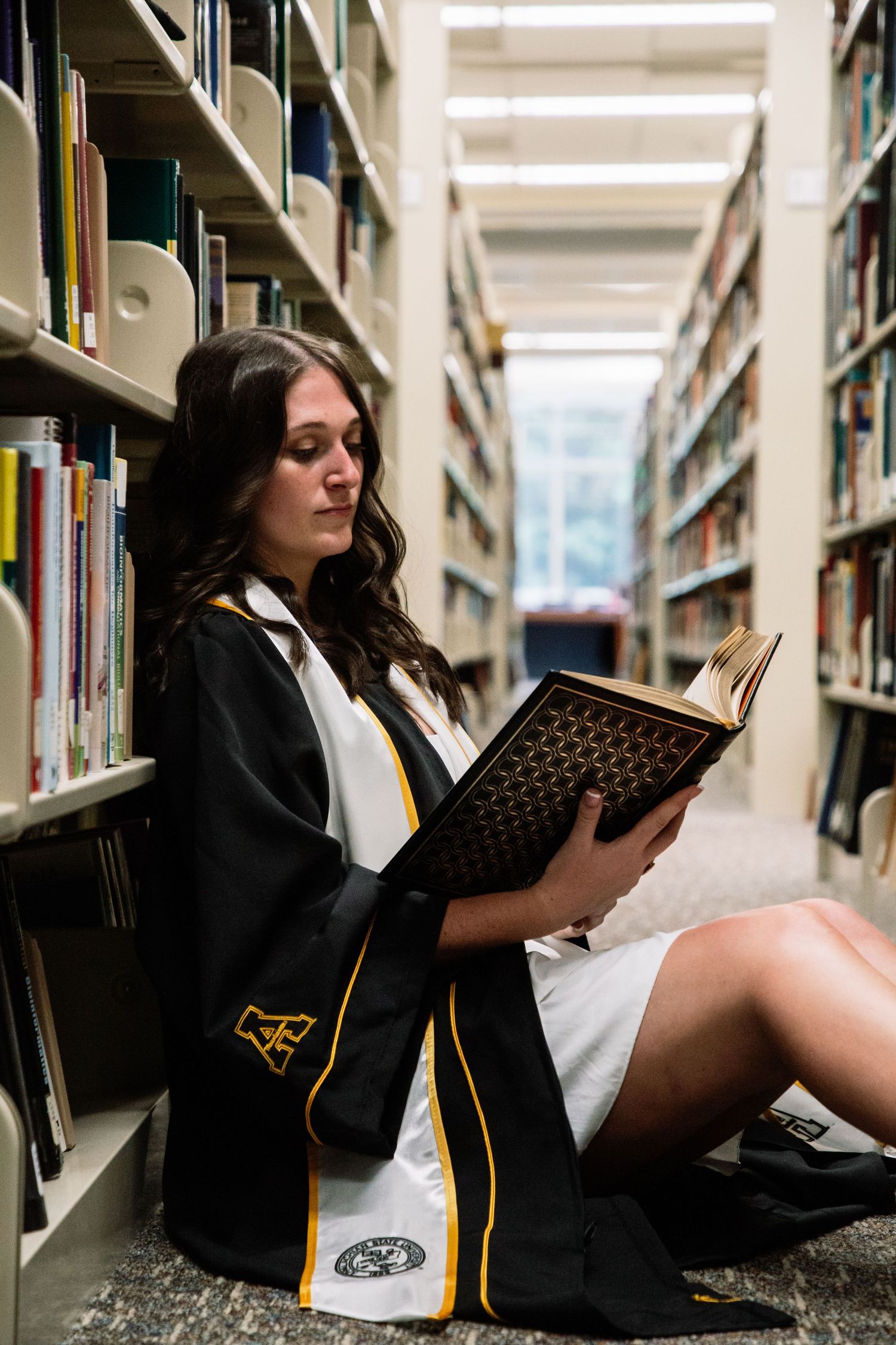 Graduate in black robe sitting on library floor reading large book.