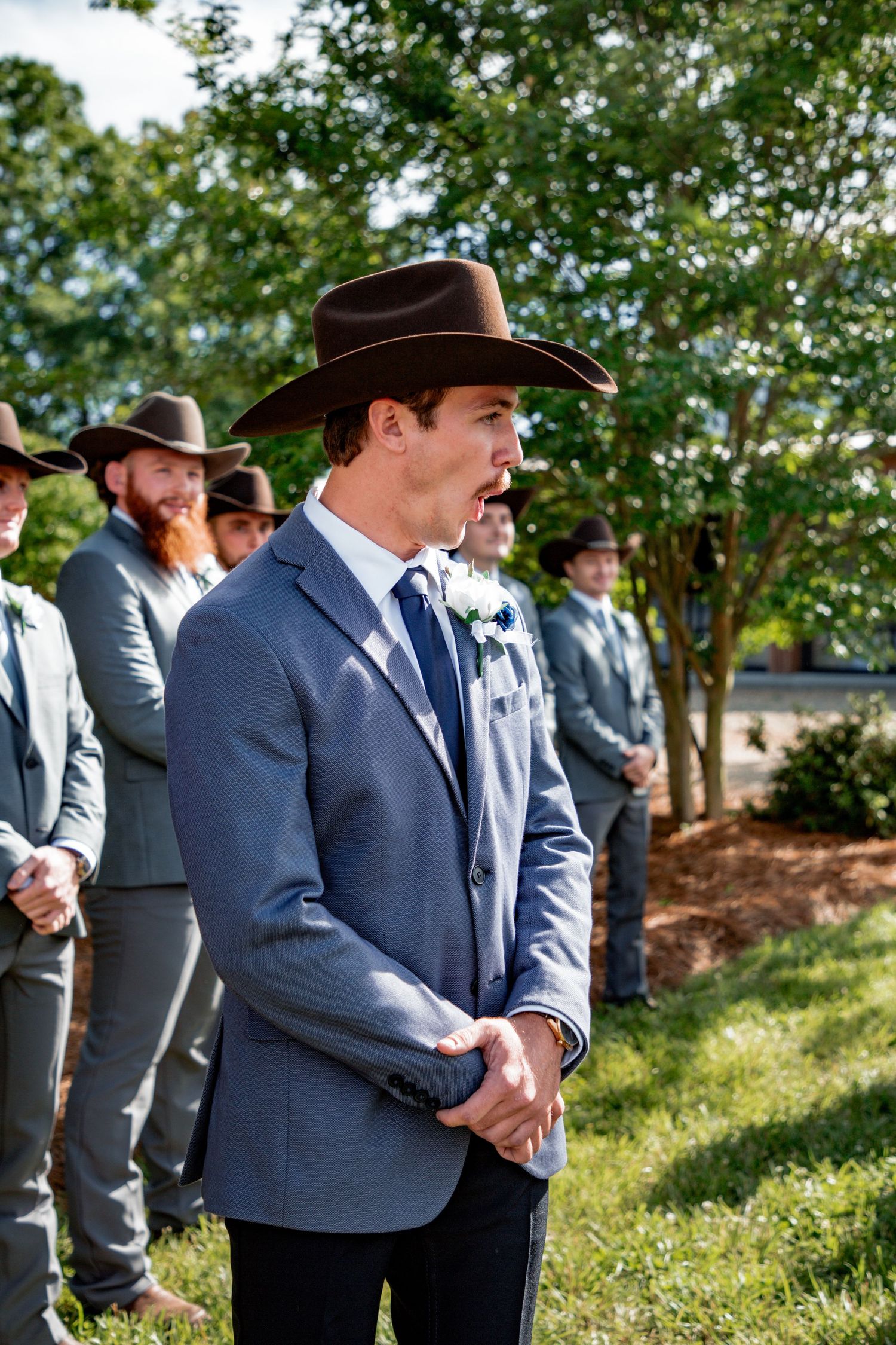 Groom in gray suit and brown cowboy hat stands with groomsmen outdoors at wedding ceremony.