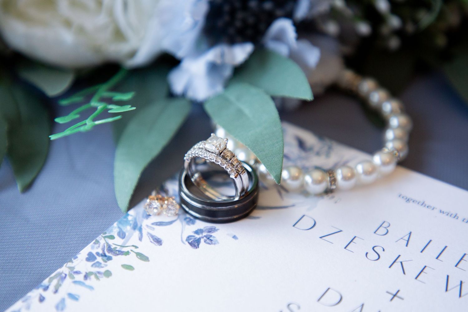 Diamond engagement ring and wedding bands resting on a floral invitation with pearl bracelet nearby.