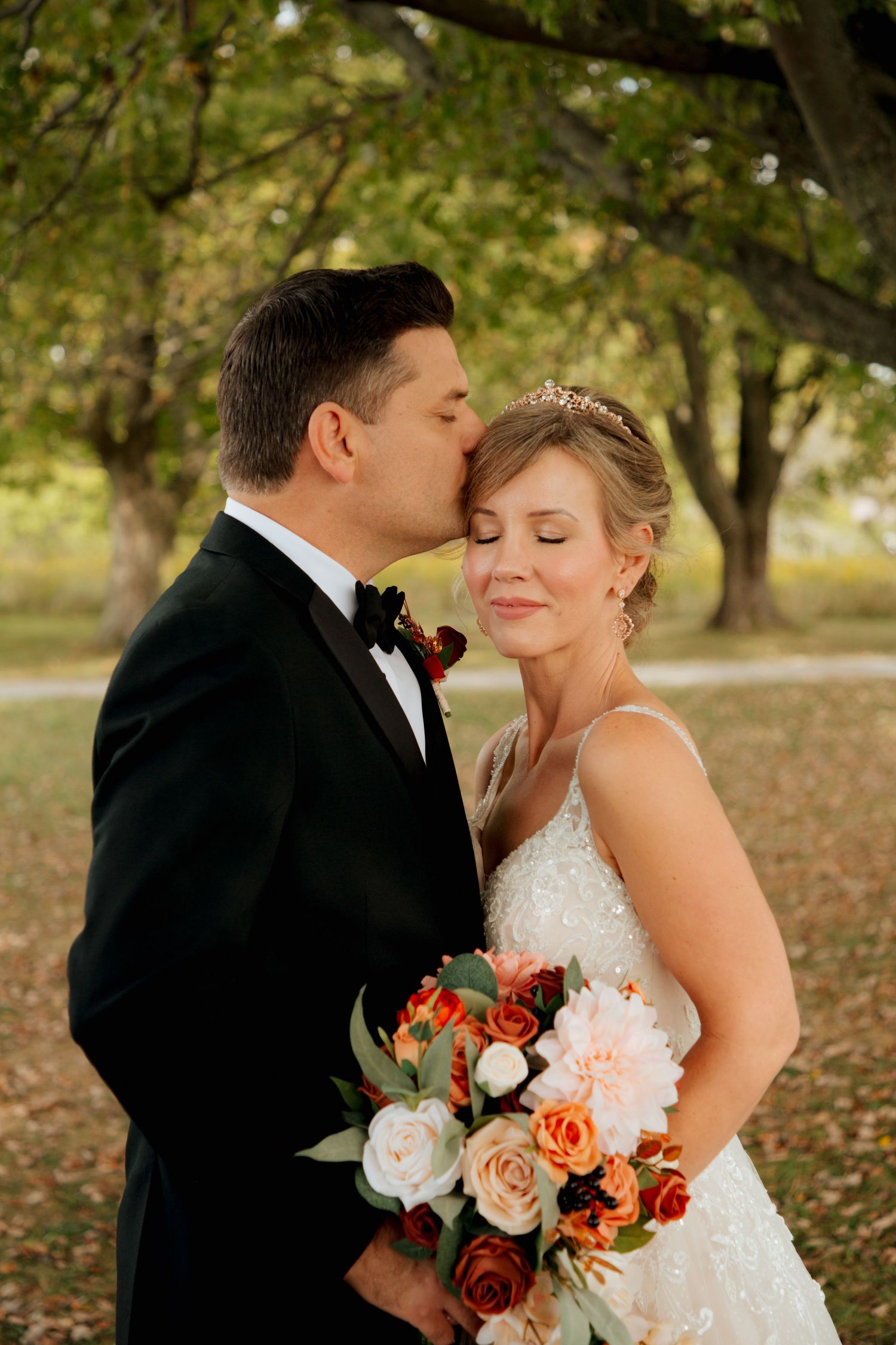 Groom in black tuxedo kisses bride on temple as she smiles holding fall bouquet.