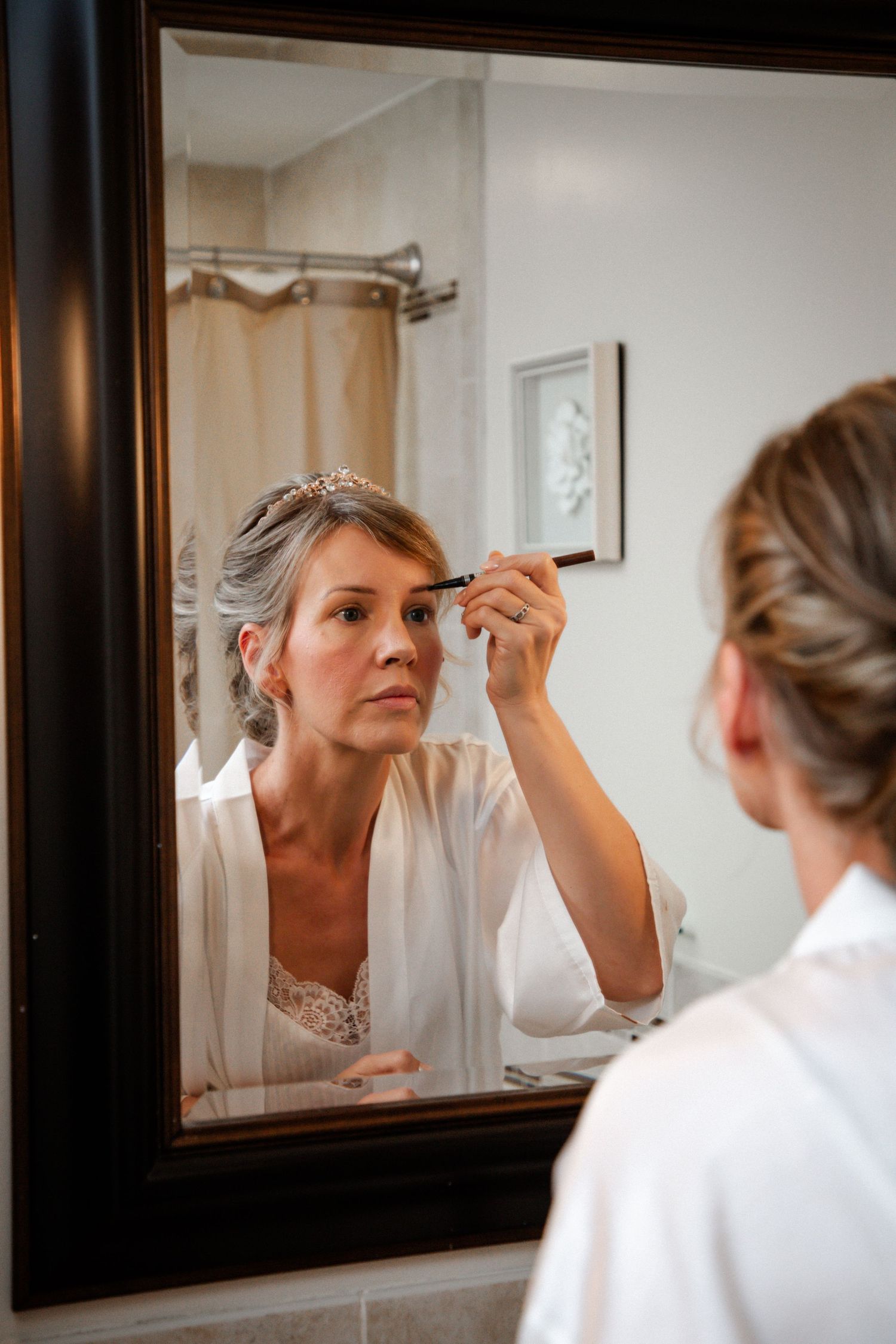 Mature bride in white robe applying makeup while looking in bathroom mirror.