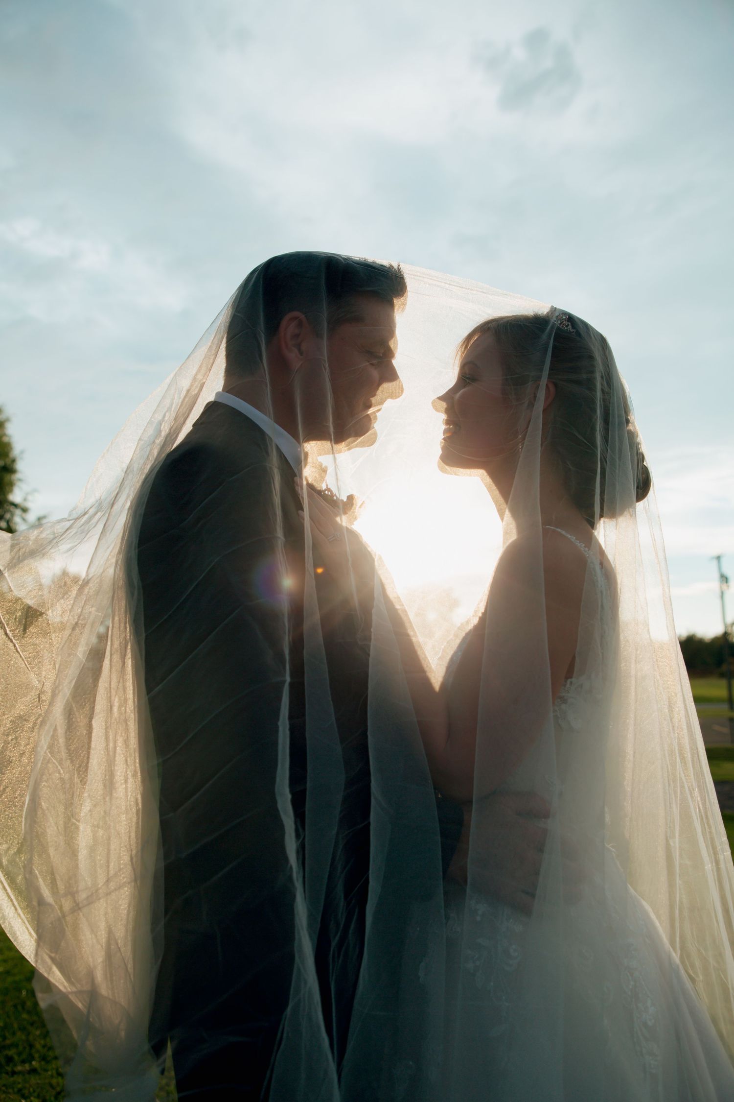 Bride and groom stand face to face beneath flowing veil with beautiful golden hour sunlight on their wedding day.