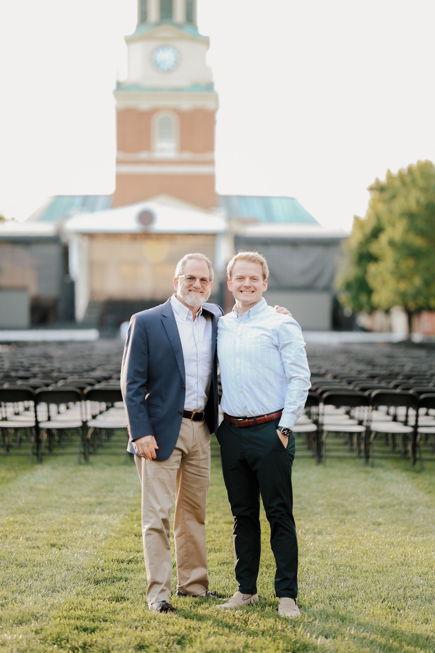 Two men smiling together on green lawn with rows of black chairs and brick clock tower behind them.