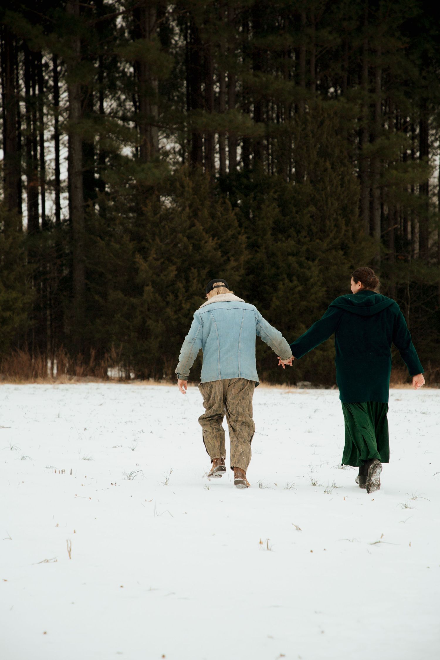 Couple holding hands walking through snowy field toward pine forest in winter.
