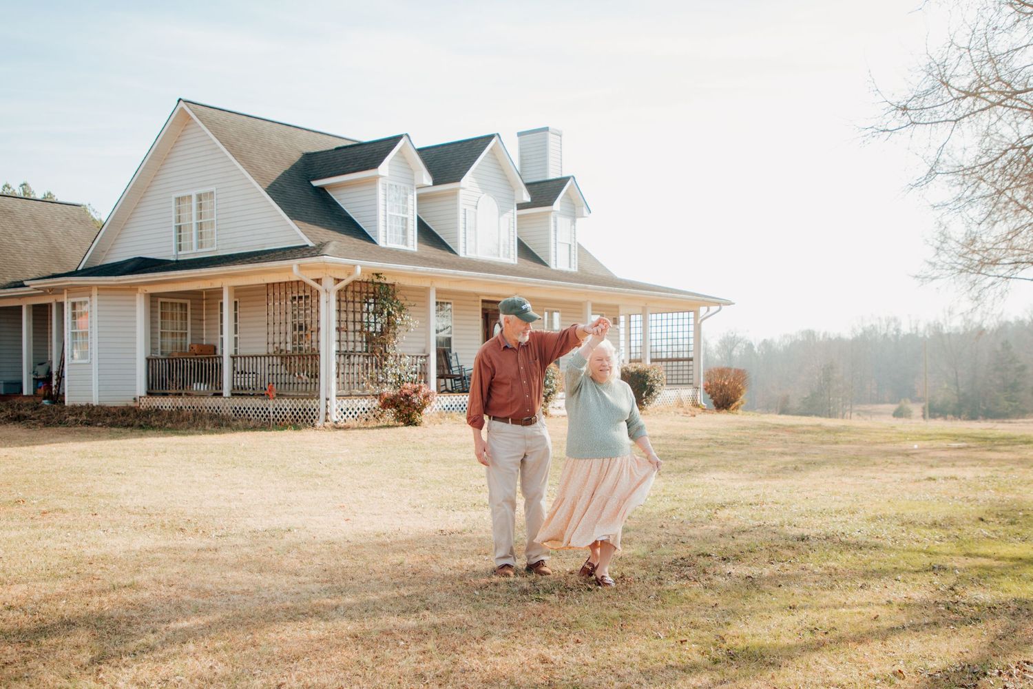Joyful senior couple dancing together in a sunny yard in front of a white farmhouse.