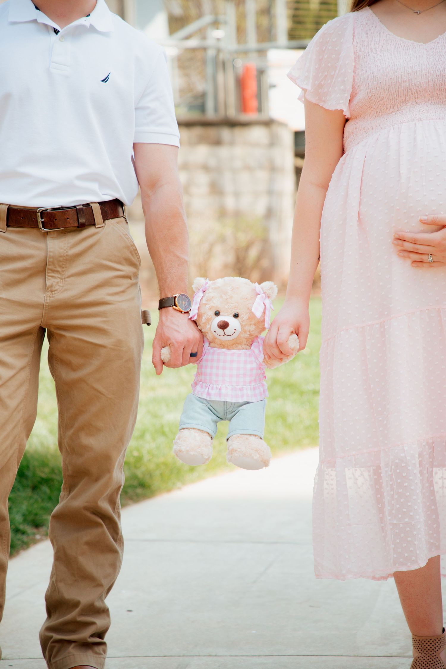 Expecting couple holding hands with a pink teddy bear between them on a walkway.