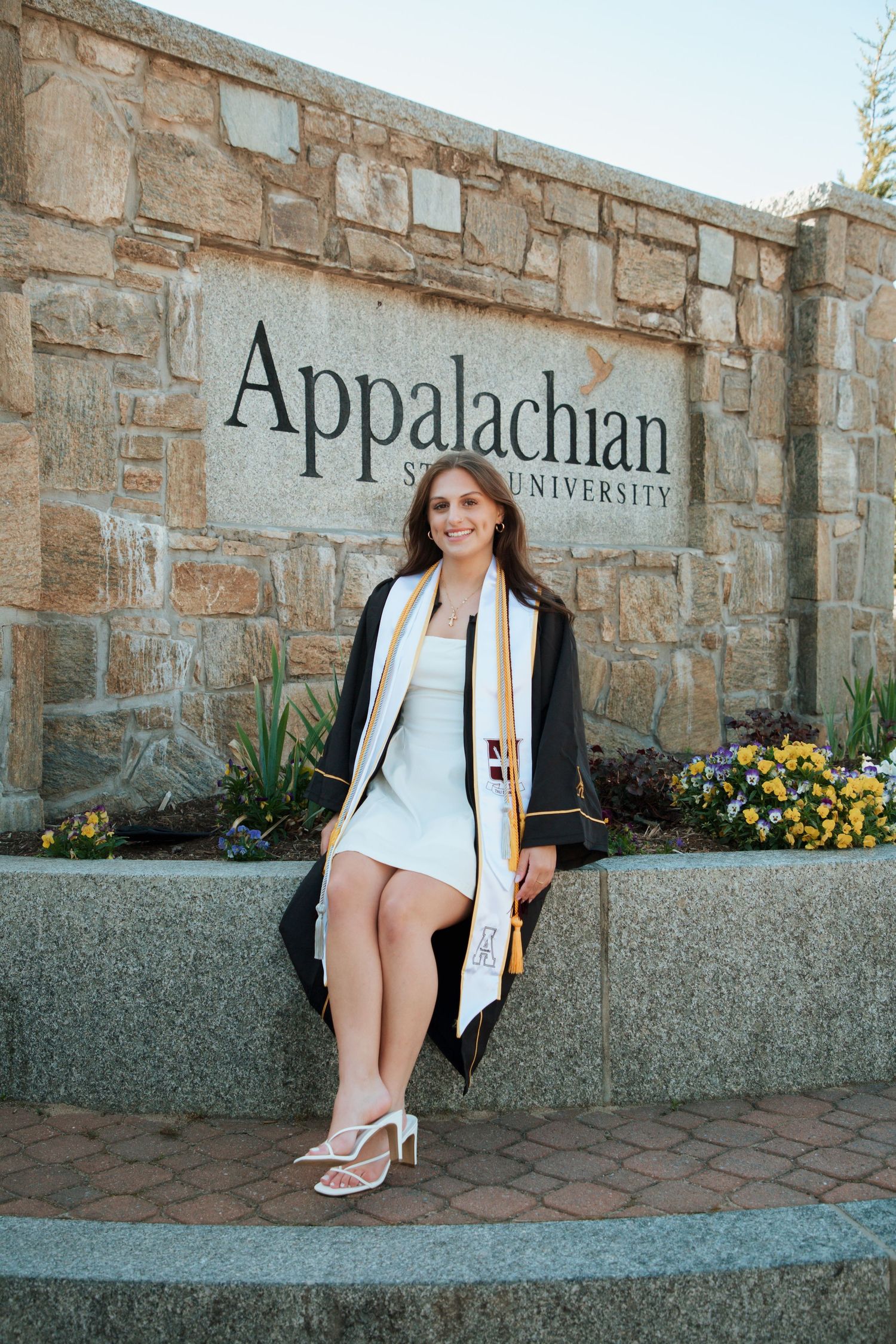 Smiling graduate in black gown sitting on ledge at Appalachian State University entrance sign.