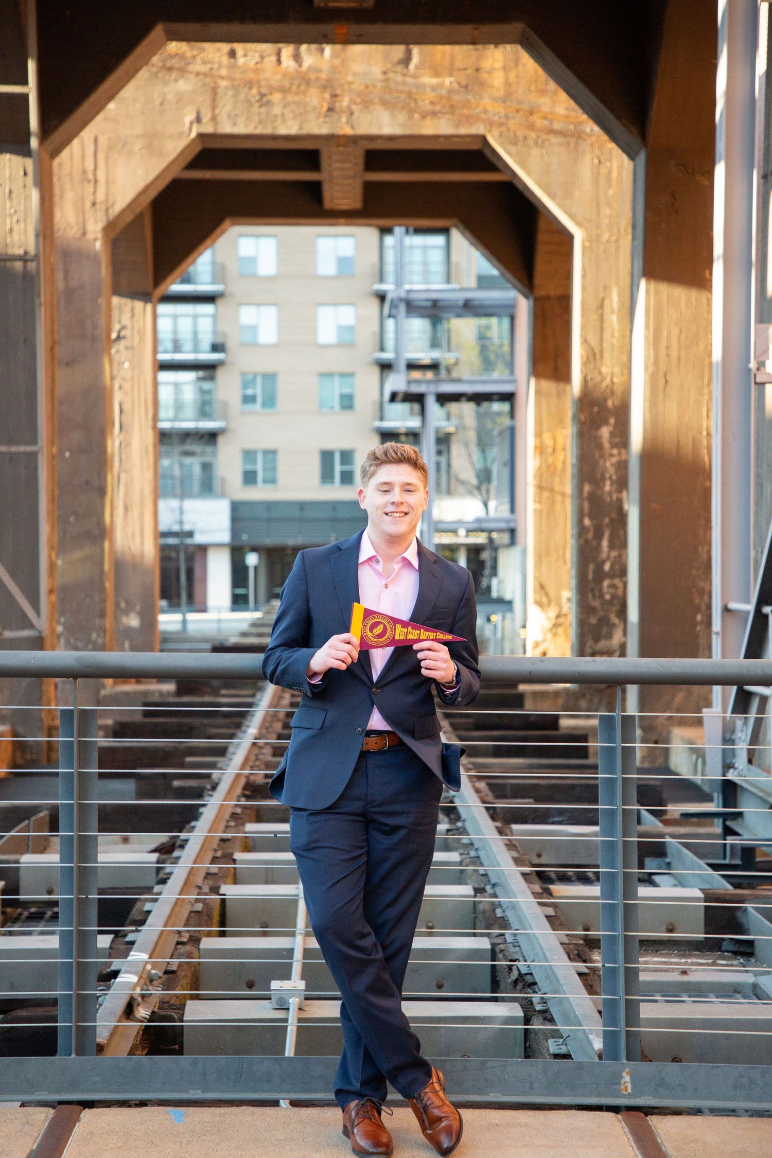 Smiling young man in navy suit holding a college pennant on an industrial bridge.