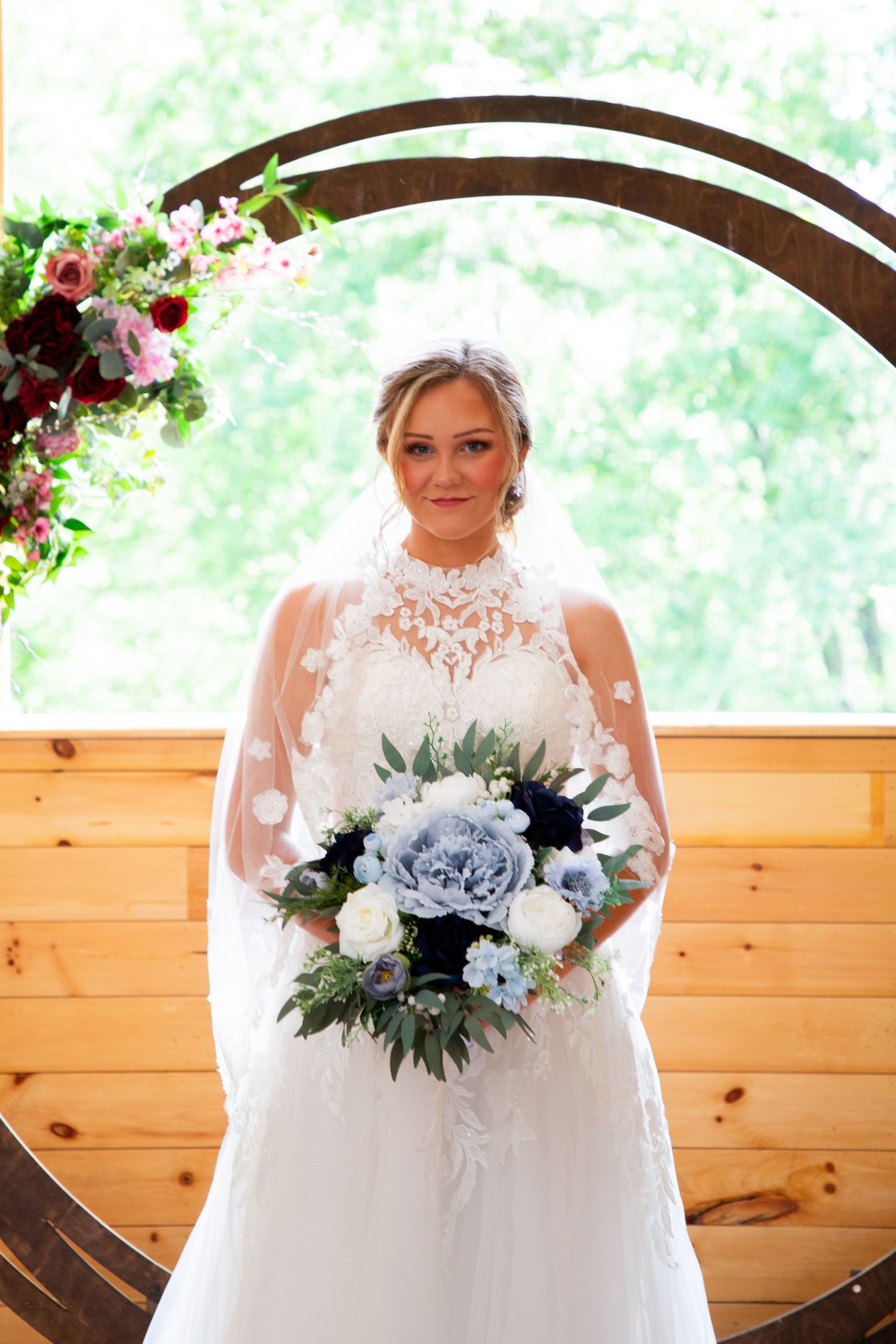Bride in lace halter gown holds blue and white bouquet under circular floral arch in rustic wood chapel.