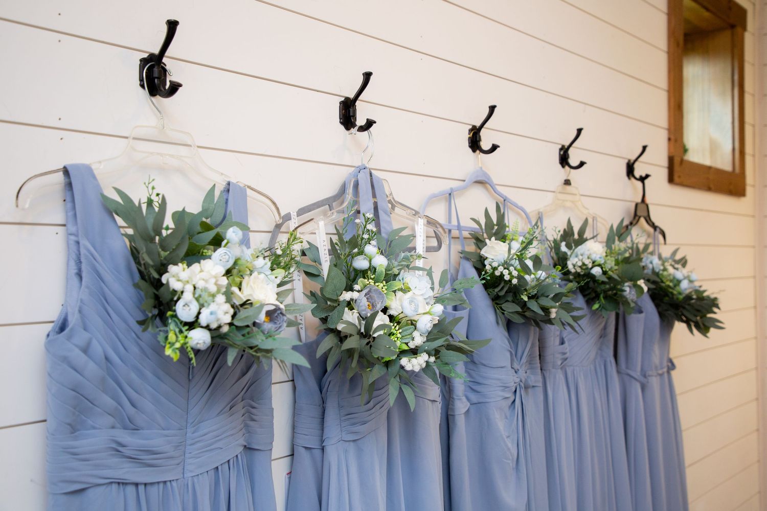 Dusty blue bridesmaid dresses hanging on hooks with white and green floral bouquets at a wedding venue.