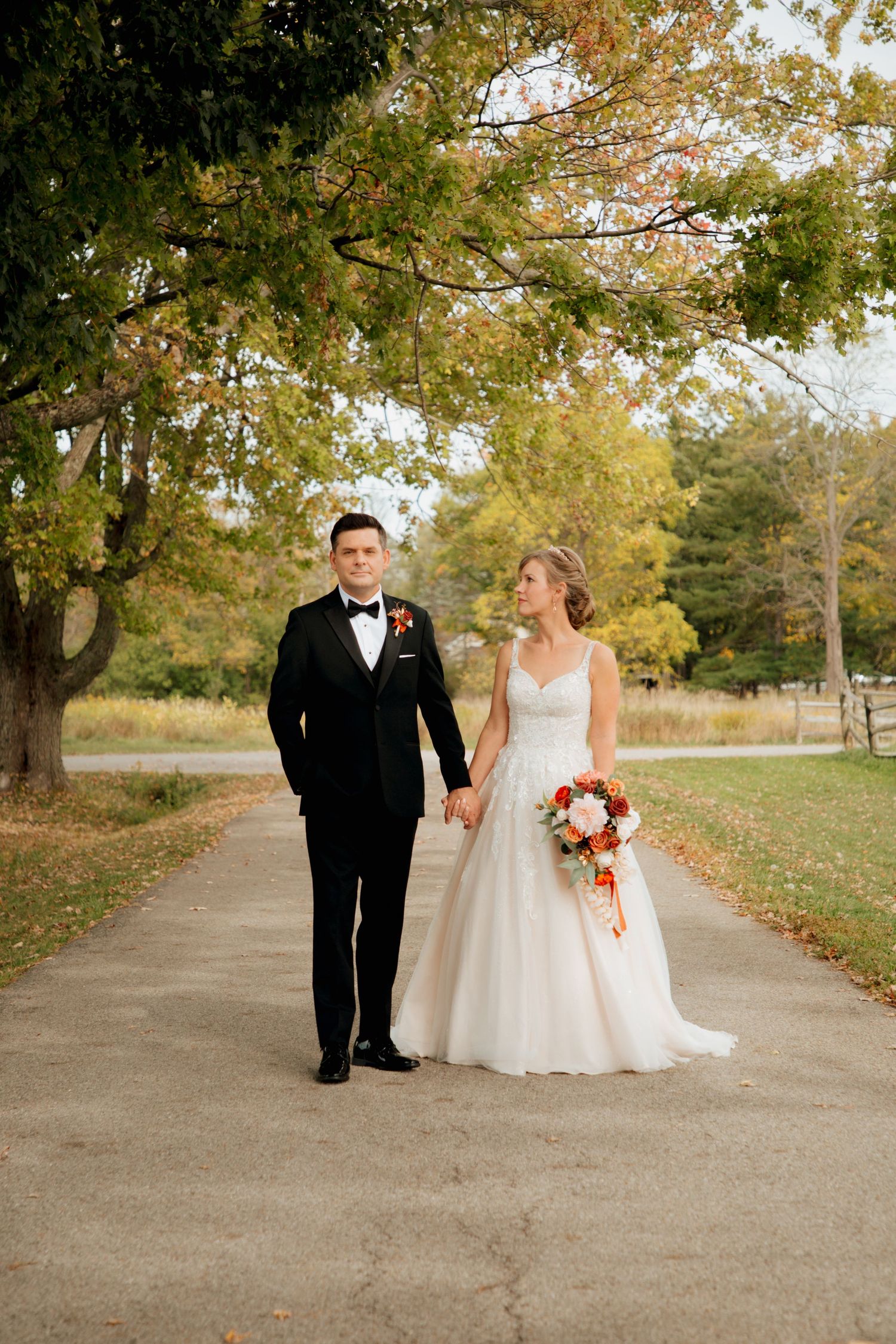 Bride and groom hold hands walking on scenic fall path with autumn foliage backdrop.