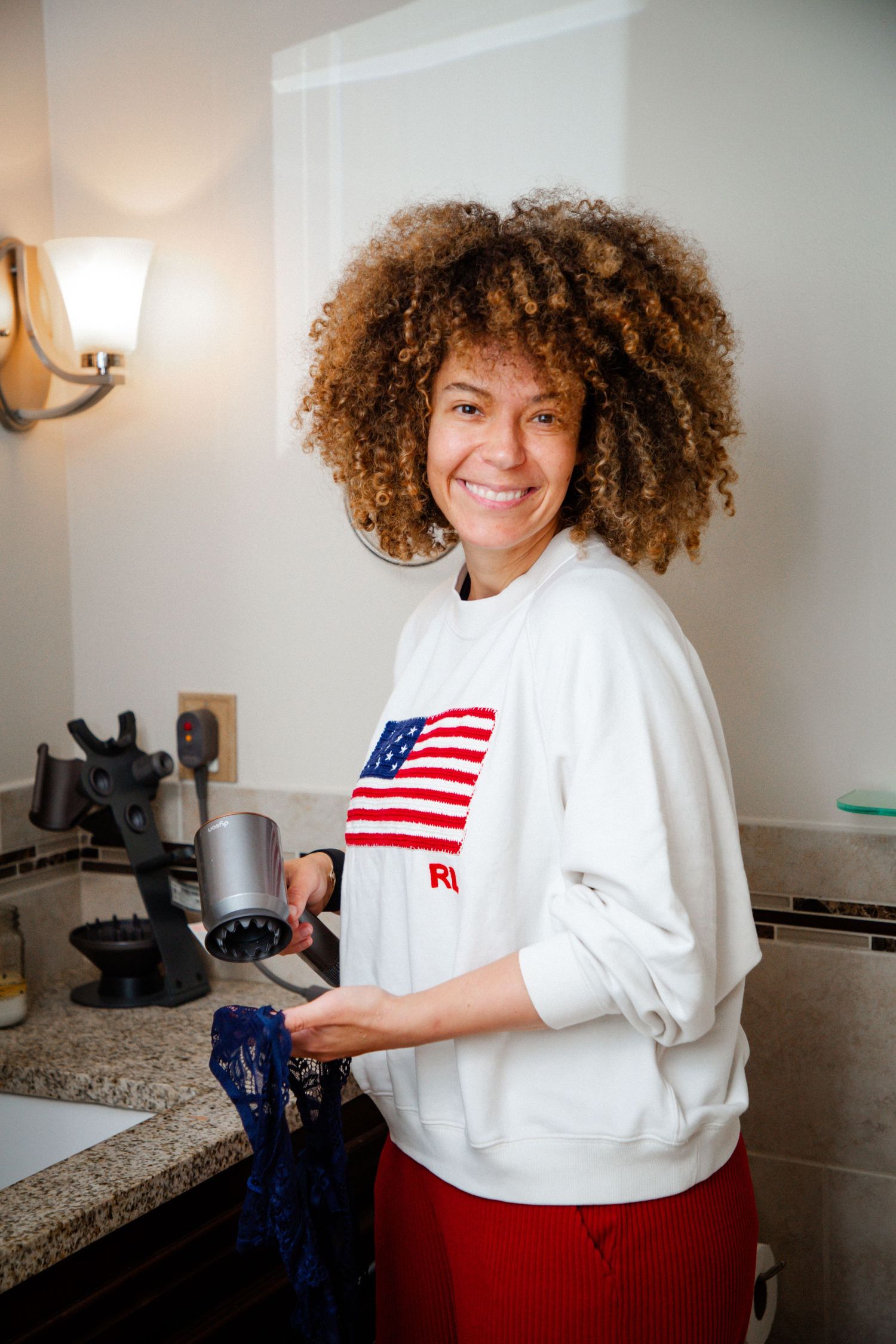 Woman in American flag sweatshirt smiling while using a milk frother in a modern kitchen.