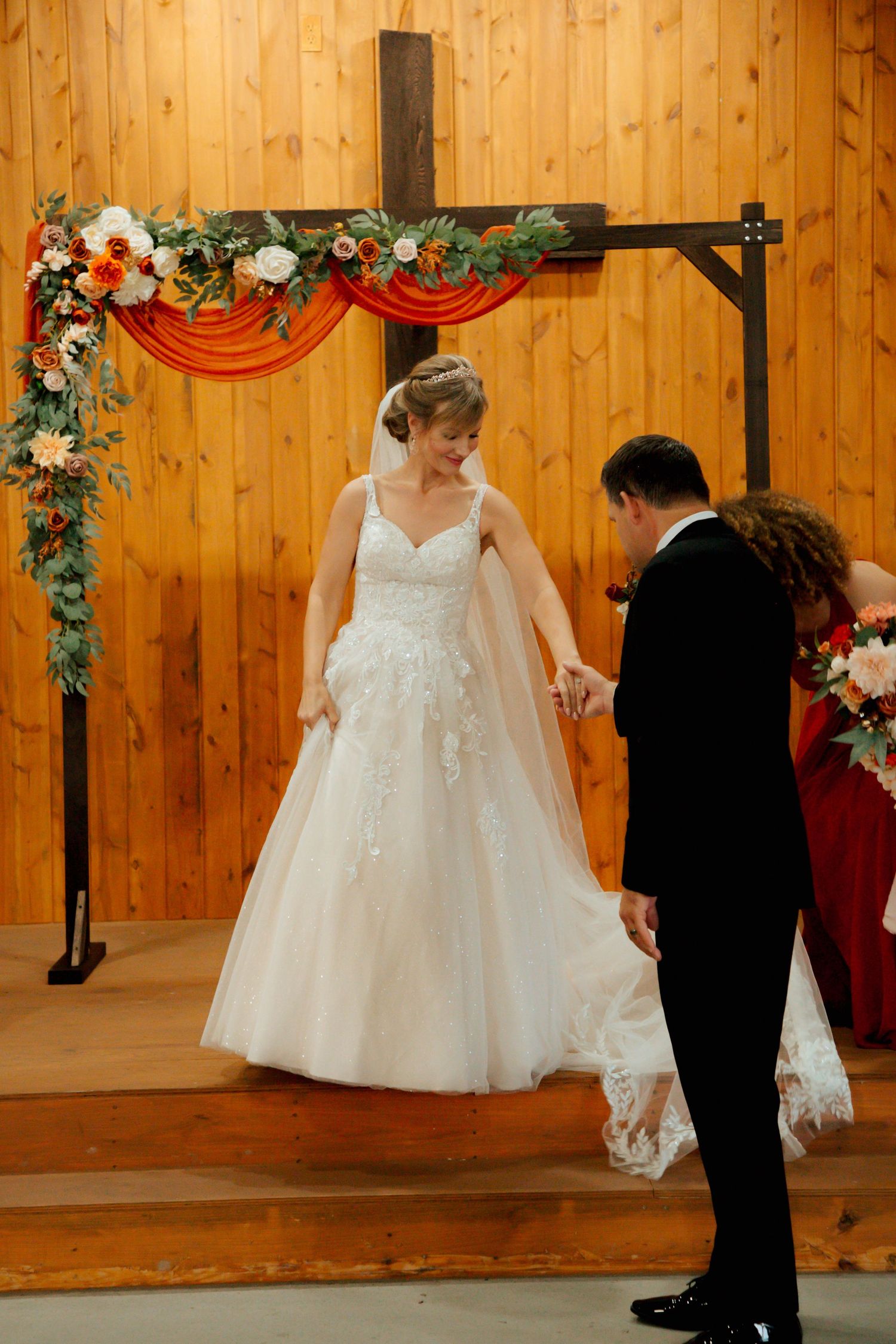 Smiling bride in lace gown holds grooms hand while stepping down from rustic chapel altar.