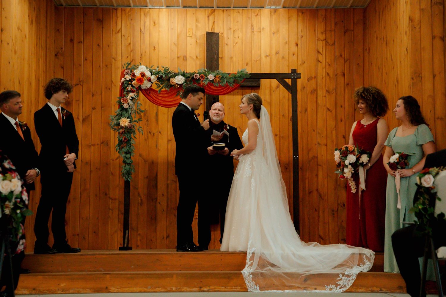 Bride in white gown and groom exchange vows beneath floral arch and wooden cross at altar.