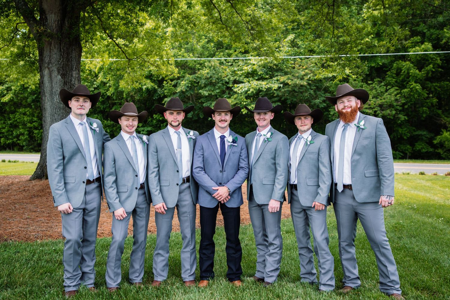 Groom and six groomsmen in gray suits and cowboy hats smile together for a formal outdoor wedding portrait.